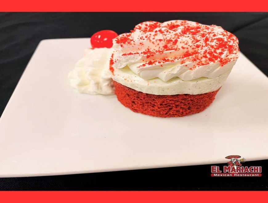 A red velvet cake topped with white frosting and red sprinkles, served with whipped cream and a cherry on a white plate.