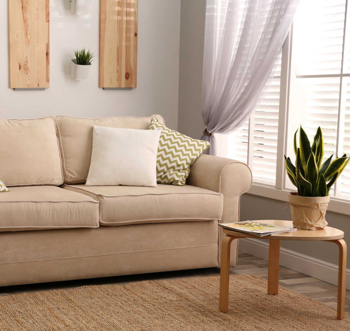 Light filled loungeroom coming through window with open venetian blinds and pulled back sheer curtains. Soft fabric couch with cushions, on mat with magazine and plant sitting on timber coffee table.