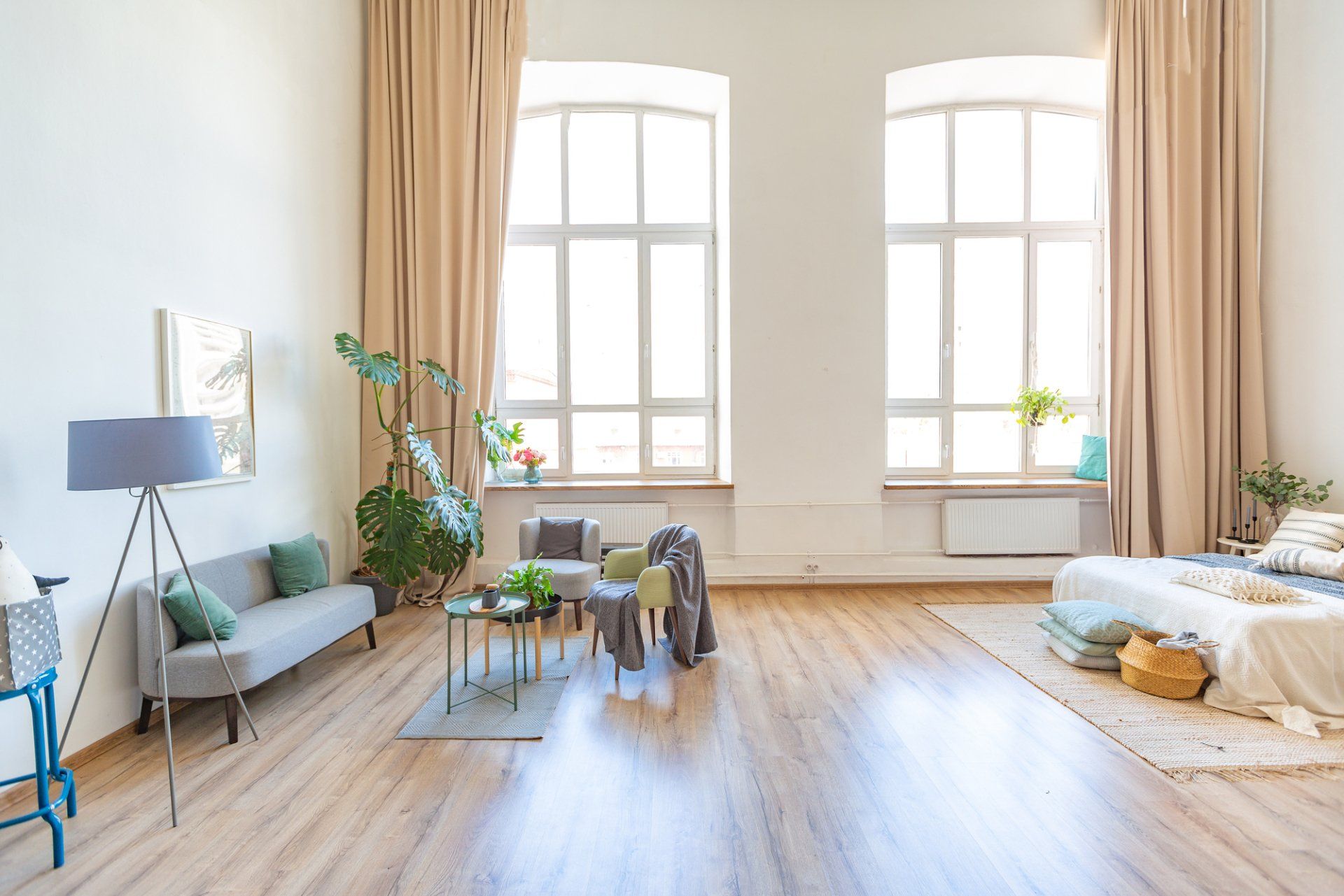 Spacious and high ceiling bedroom with timber floors, bed on mat, small couch and chairs around small table. Backed by floor to ceiling drapes pulled aside to show light flowing through windows.