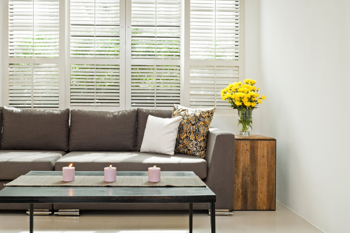 Semi open Plantation Shutters in lounge room with leather couch and cushions, coffee table with 3 pink candles, timber side table with bunch of yellow flowers in vase.