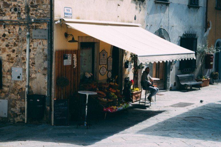 Awning extended out over road on stone corner store with potted flowers, and man sitting on stool.