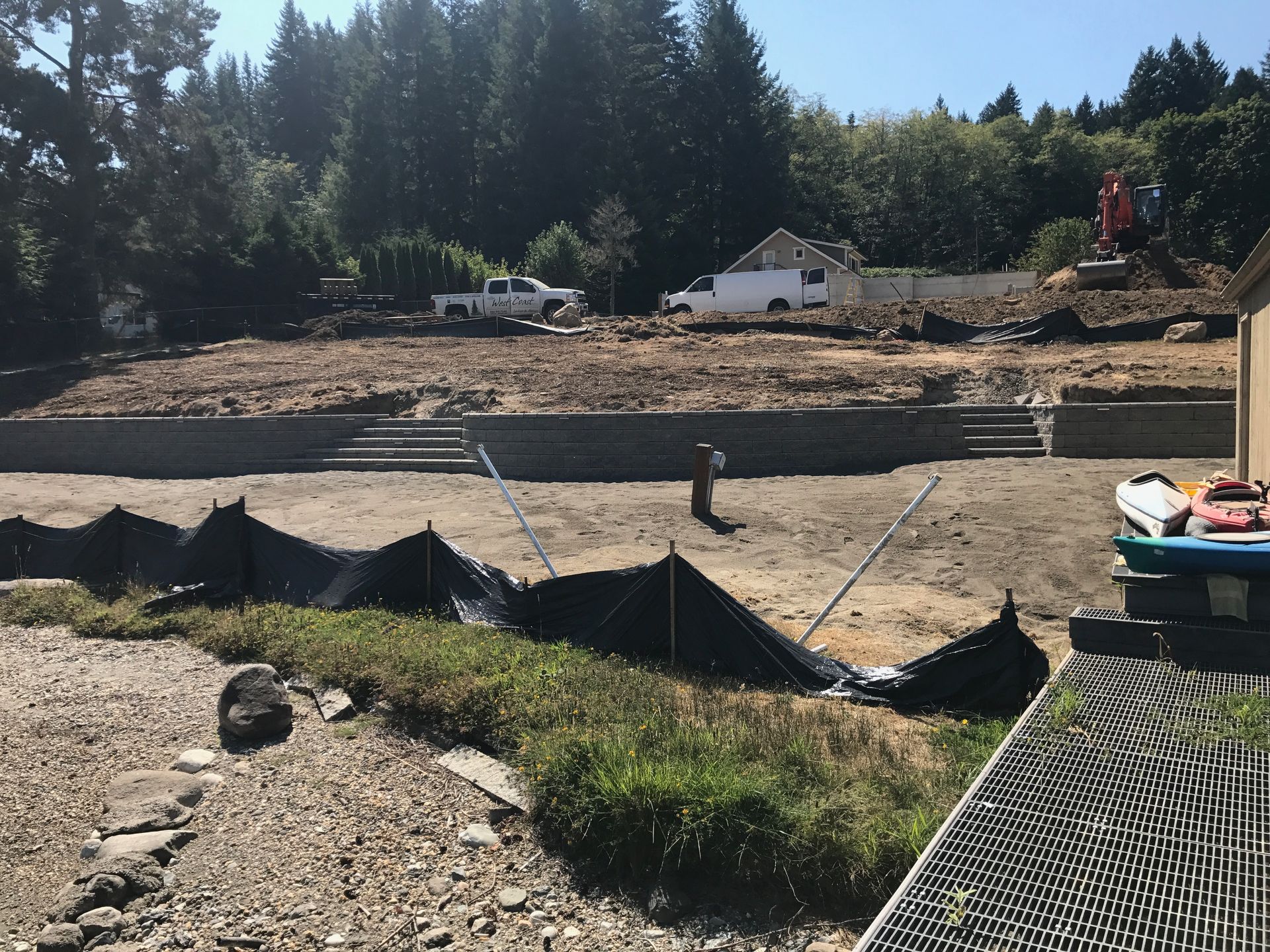 Construction site with cleared earth, black silt fence, gravel foreground, and trees behind a white house.