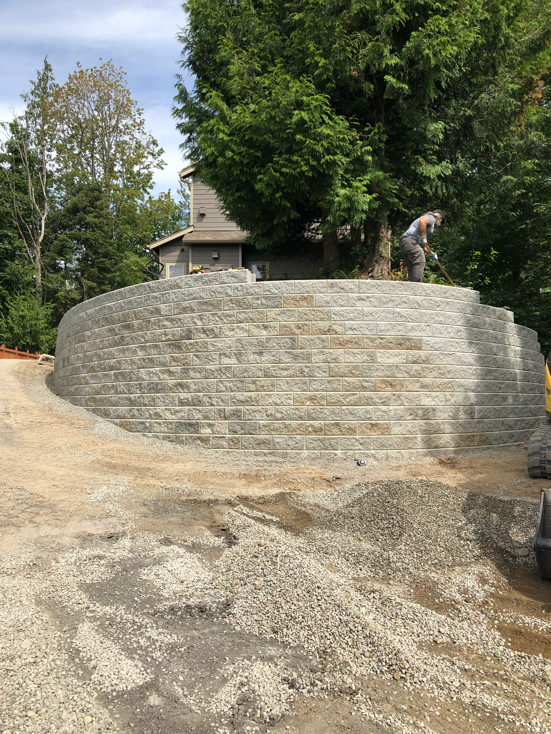A person stands on a large stone wall in a wooded outdoor area near a small building.