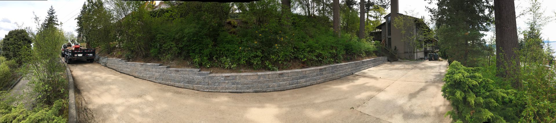 Curved stone wall beside a dirt path in a leafy garden area, with a parked vehicle on the left and trees behind.
