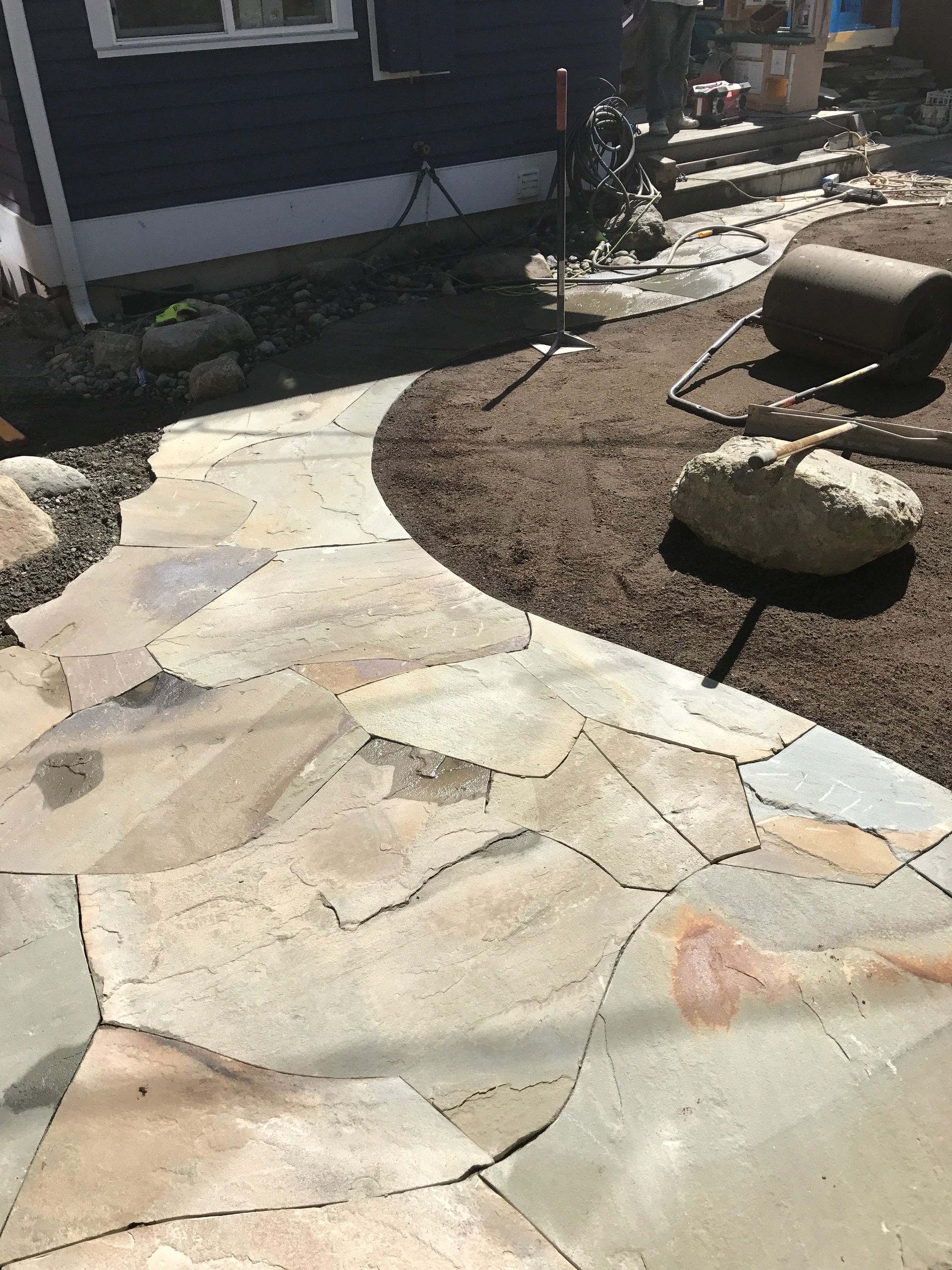 Stone patio walkway beside a house, bordered by landscaping rocks and a dirt garden bed.