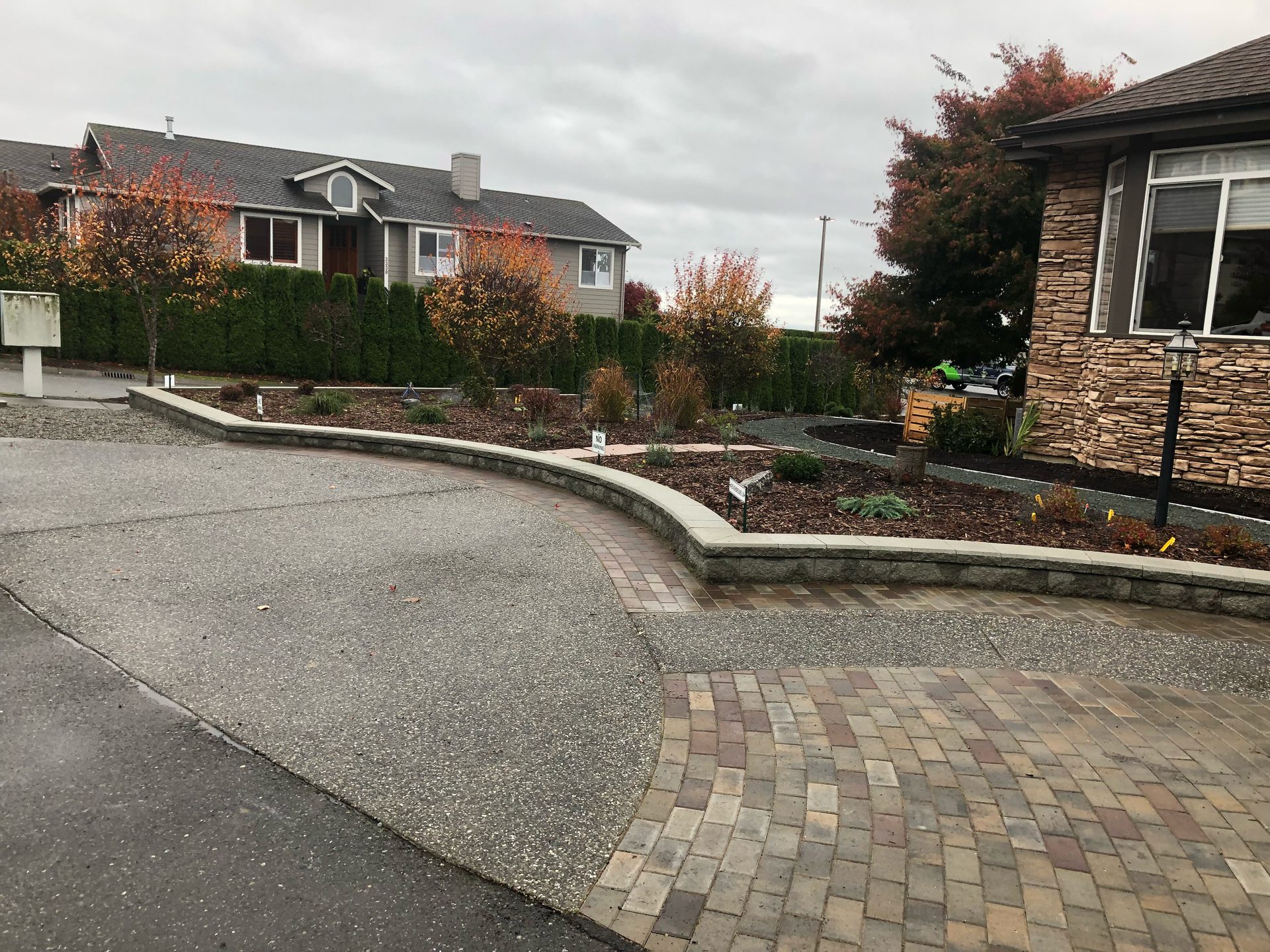 Residential street with curved curb, paved driveway, and landscaped garden beds beside houses on a cloudy day
