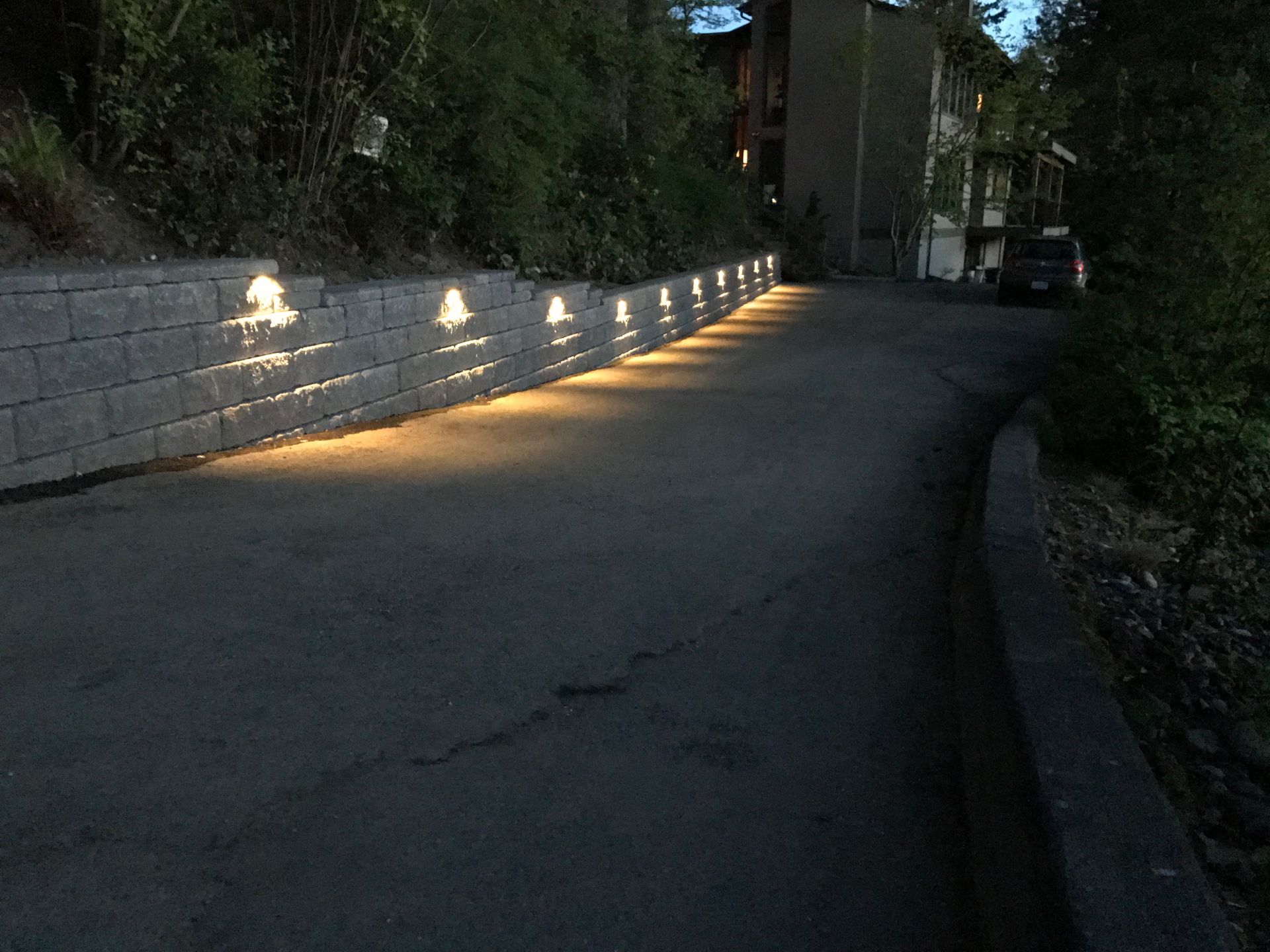 Dim driveway beside a stone wall lit by small ground lights at dusk