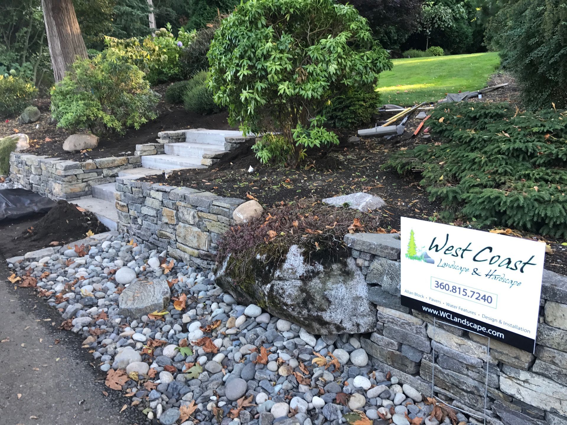 Landscape sign in a rock garden with stone steps, shrubs, and a pebble-lined path.