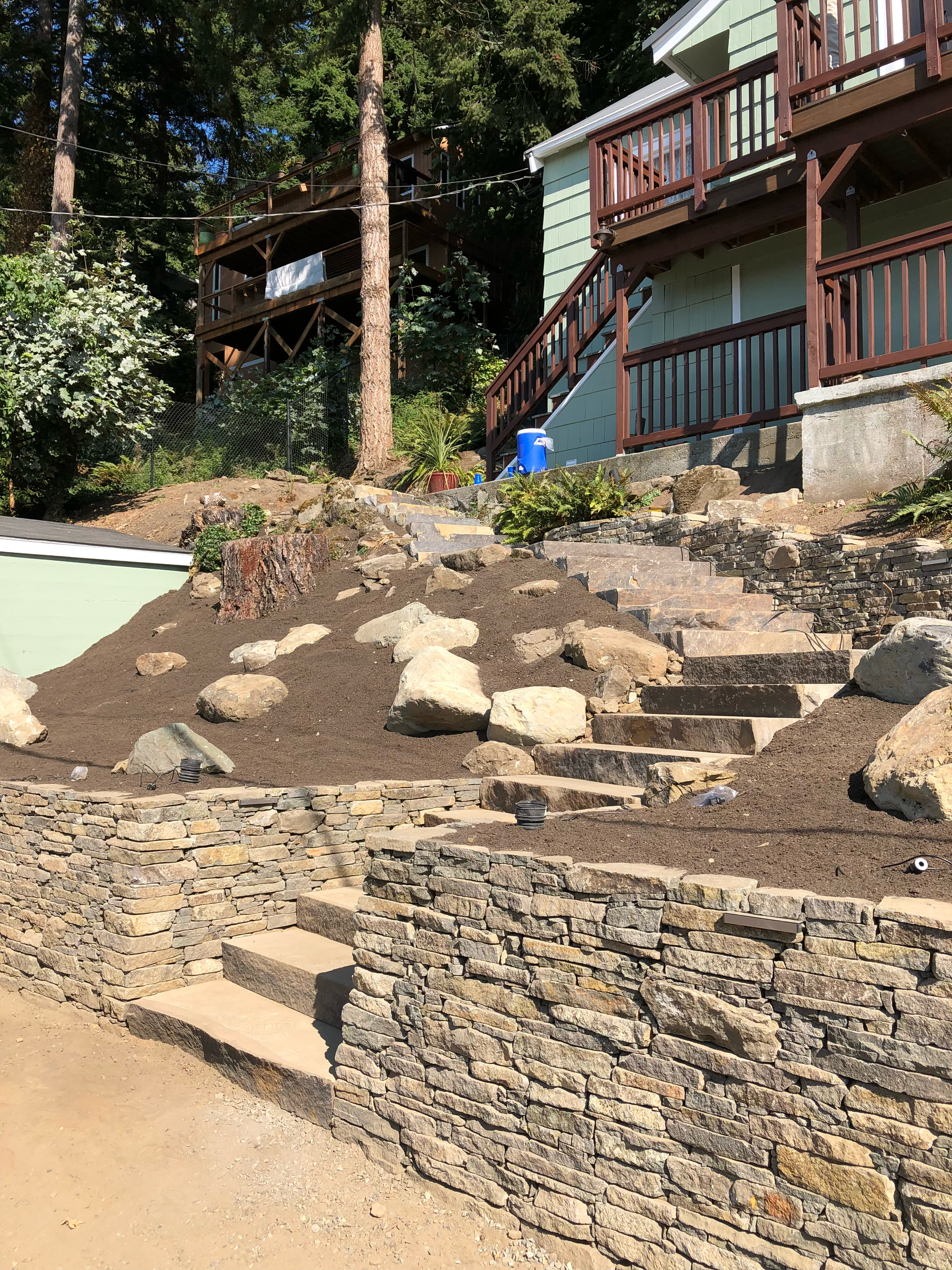 Outdoor sloped landscaping with stone retaining walls, steps, and a house deck in the background