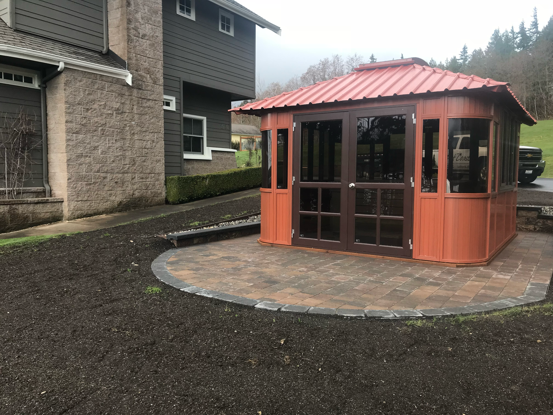 Small orange gazebo with red roof on a paved patio beside a stone building in a rainy courtyard