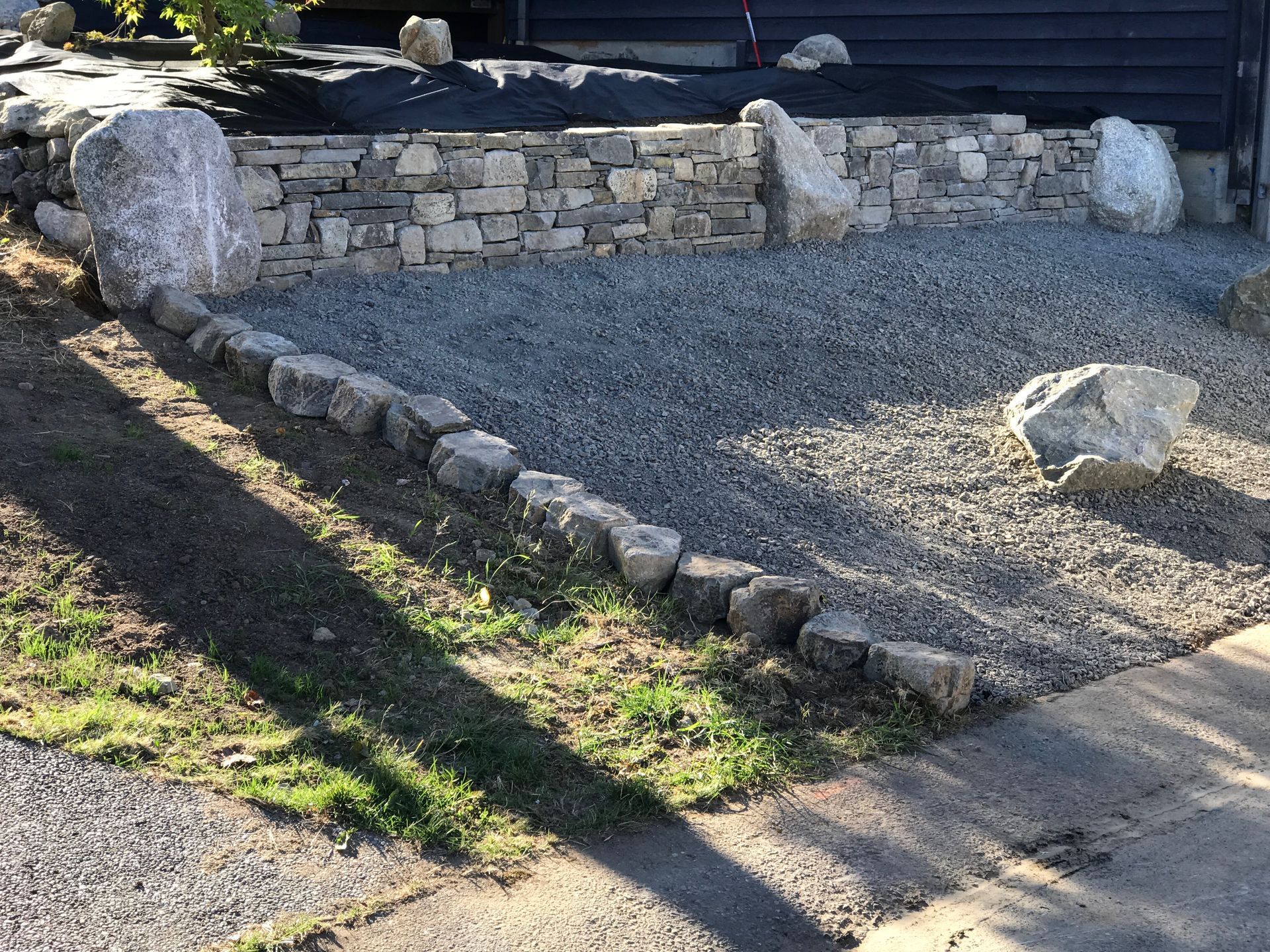 Stone gravel landscape with a low retaining wall and a large rock beside a sidewalk.