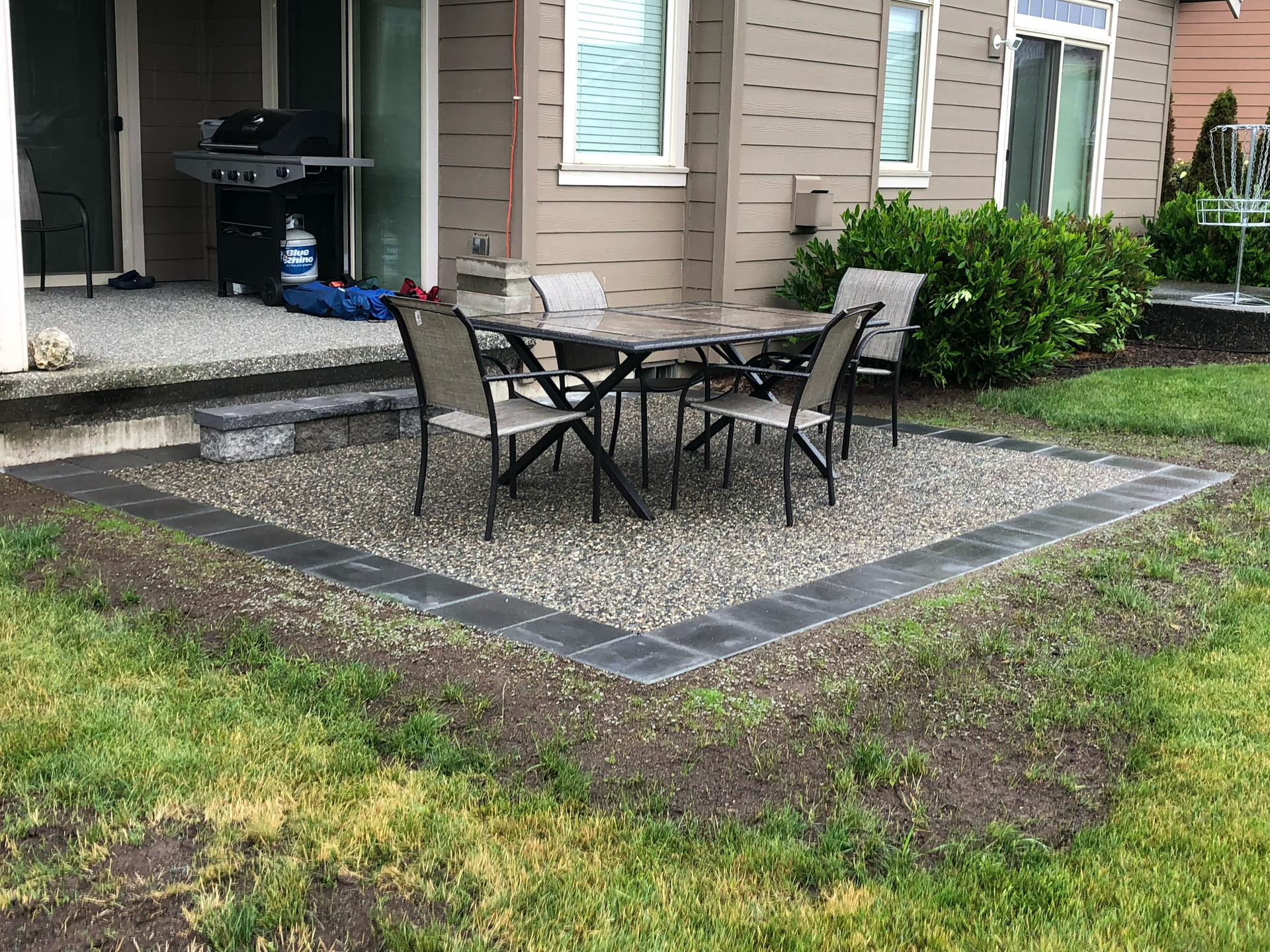 Patio with black metal table and chairs on gravel beside a house porch and grill