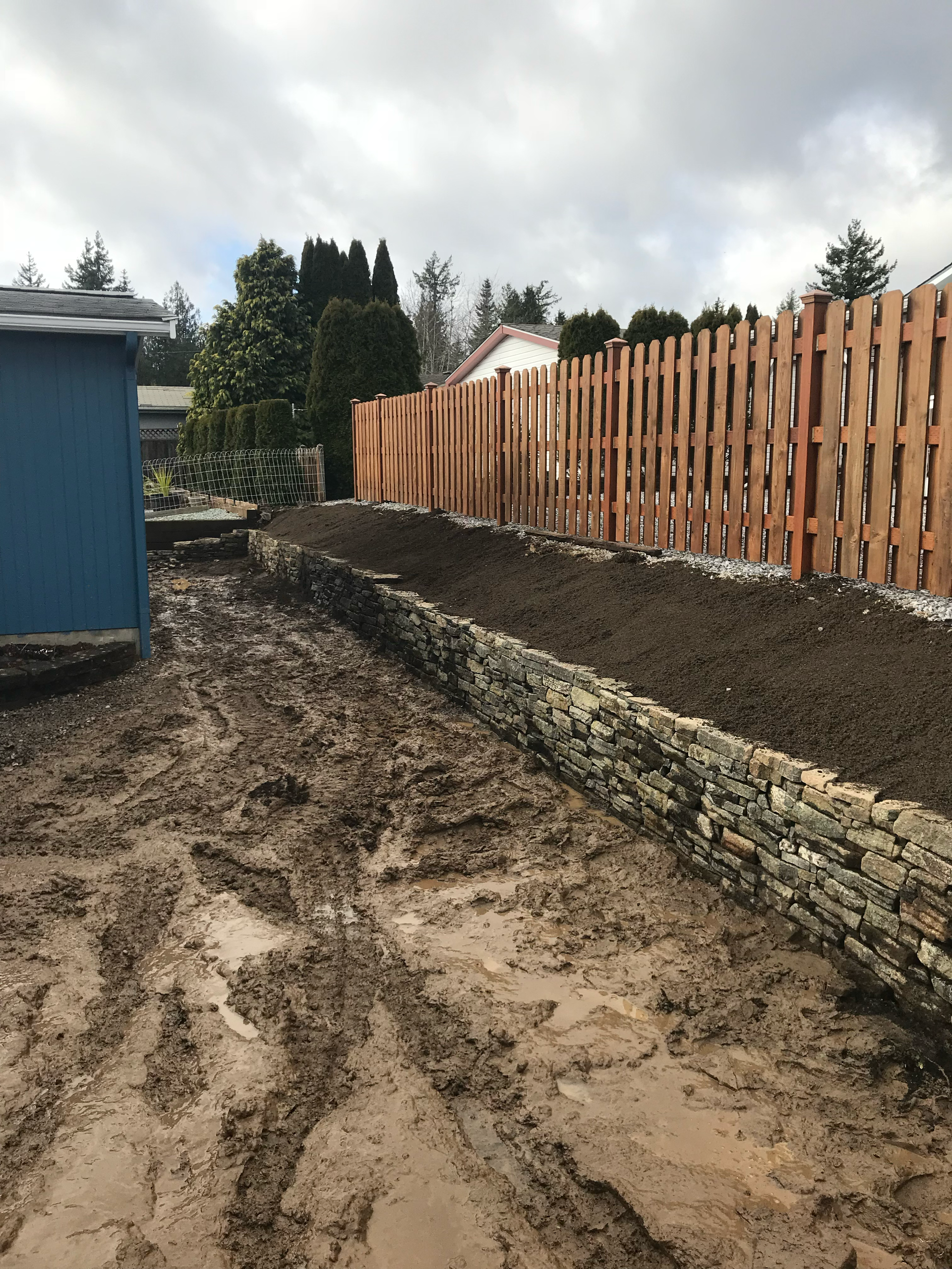 Mud-filled backyard beside a blue shed, stone retaining wall, and wooden fence under cloudy skies