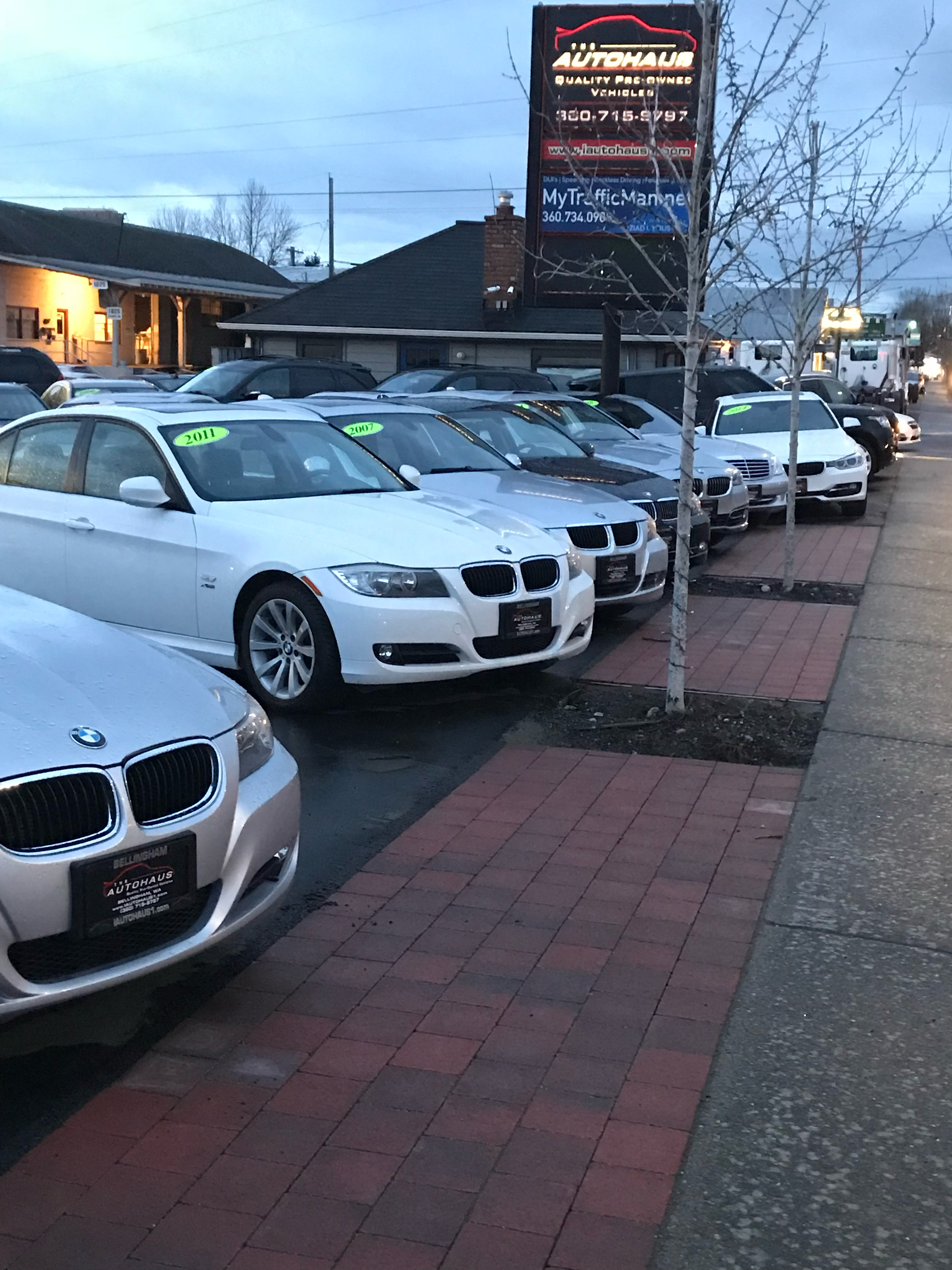 Row of parked cars along a brick sidewalk at dusk outside a lit dealership sign.