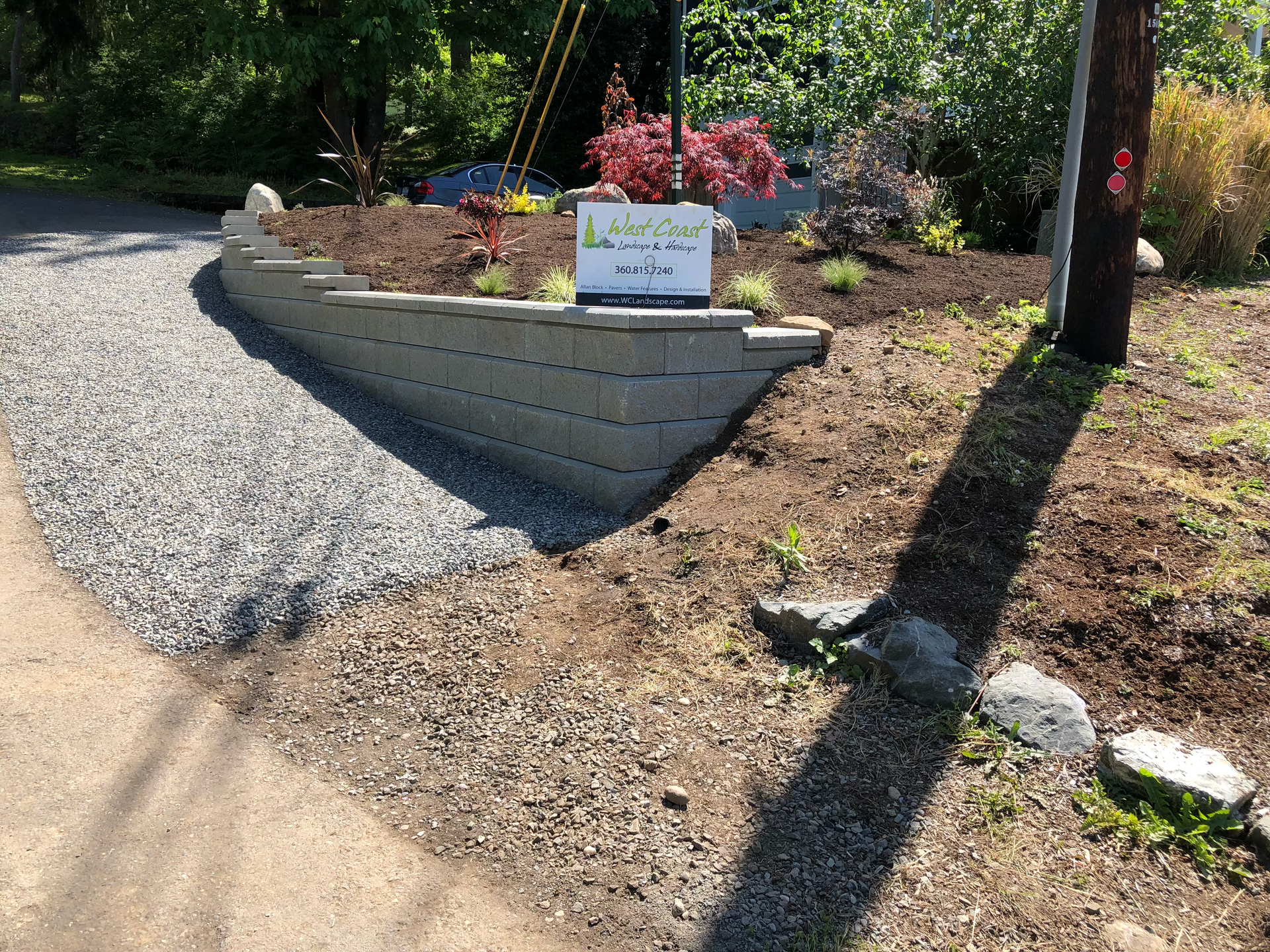 Gravel driveway beside a landscaped stone retaining wall with flowers and a small sign