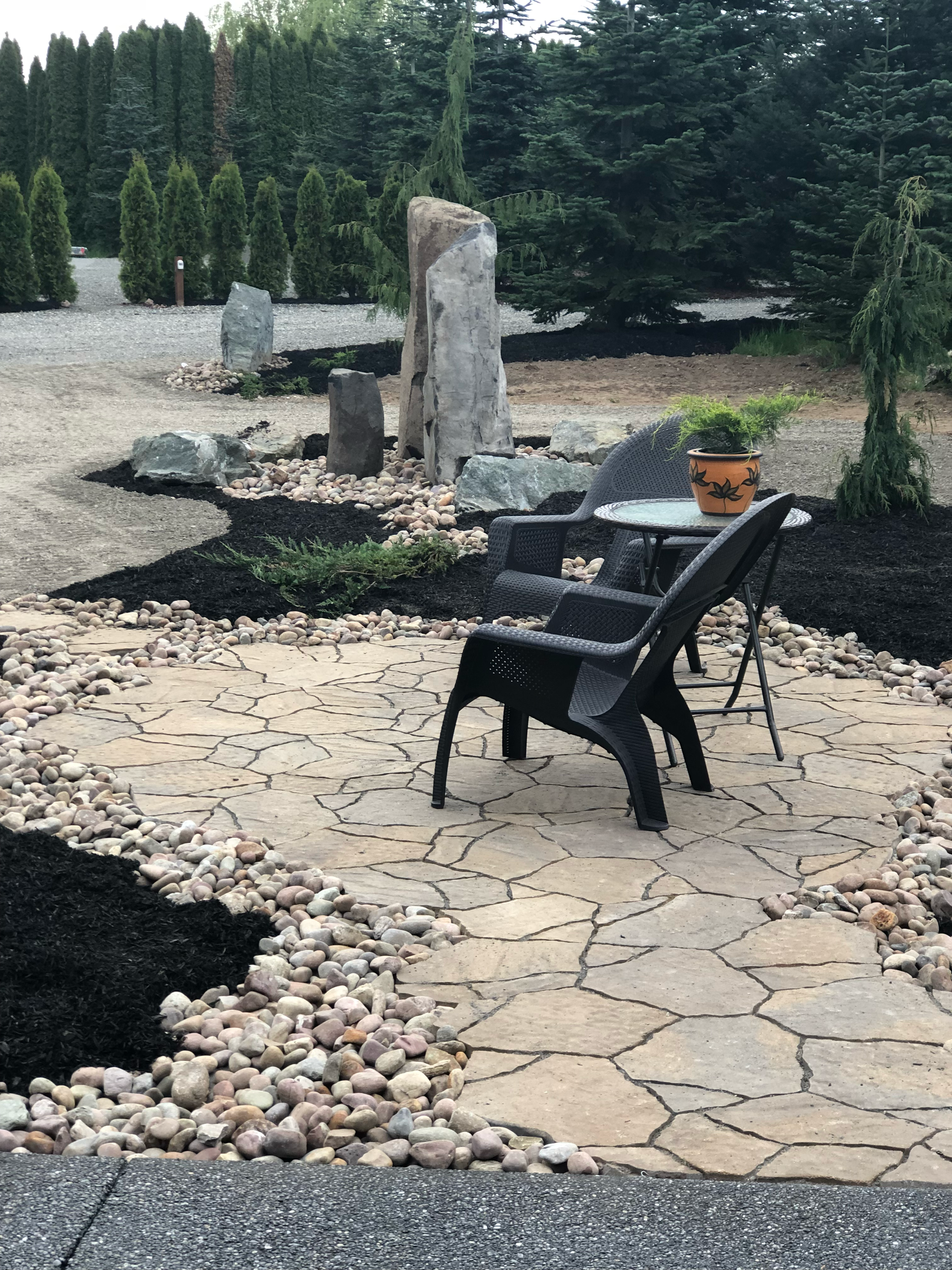 Backyard patio with a black chair, stone path, decorative rocks, and a small fountain among evergreens