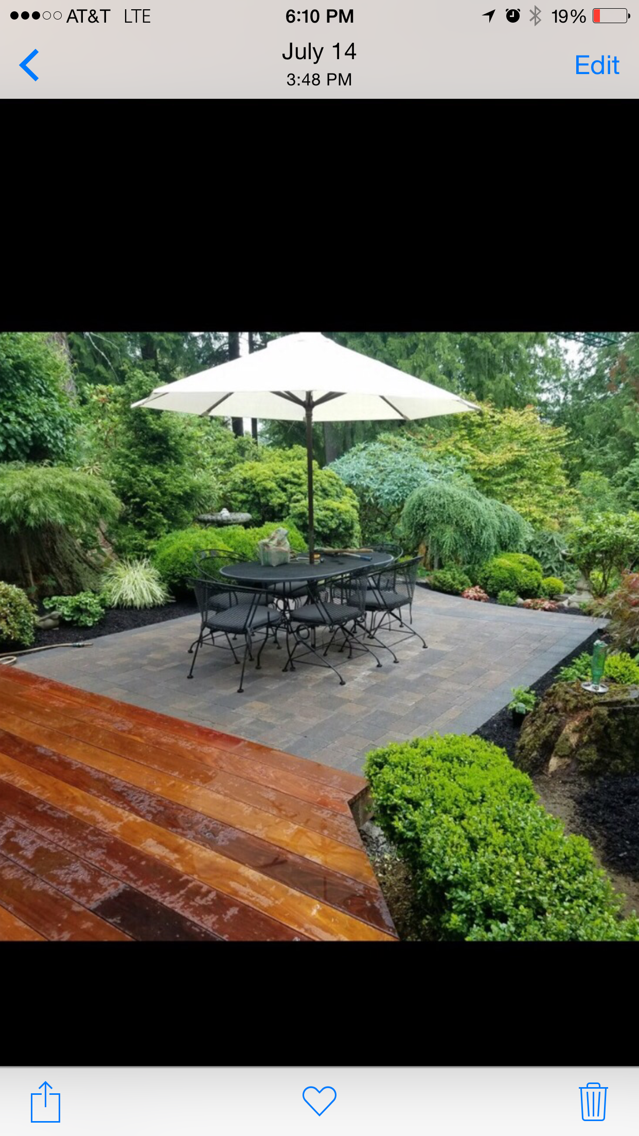 Garden patio with stone table, black chairs, and a white umbrella beside lush greenery.