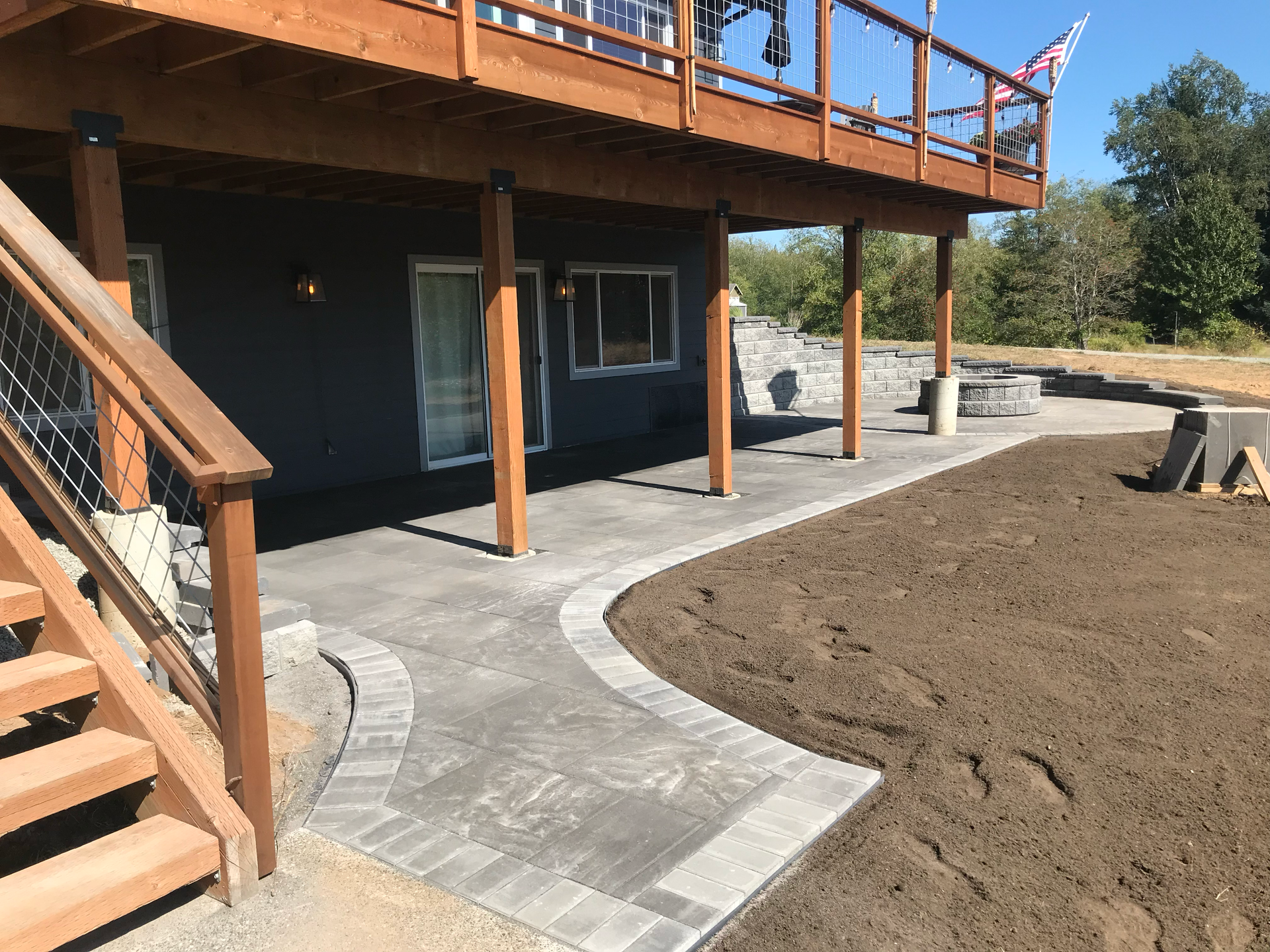 Curved concrete walkway beside a dark house with wooden deck stairs and bare soil landscaping