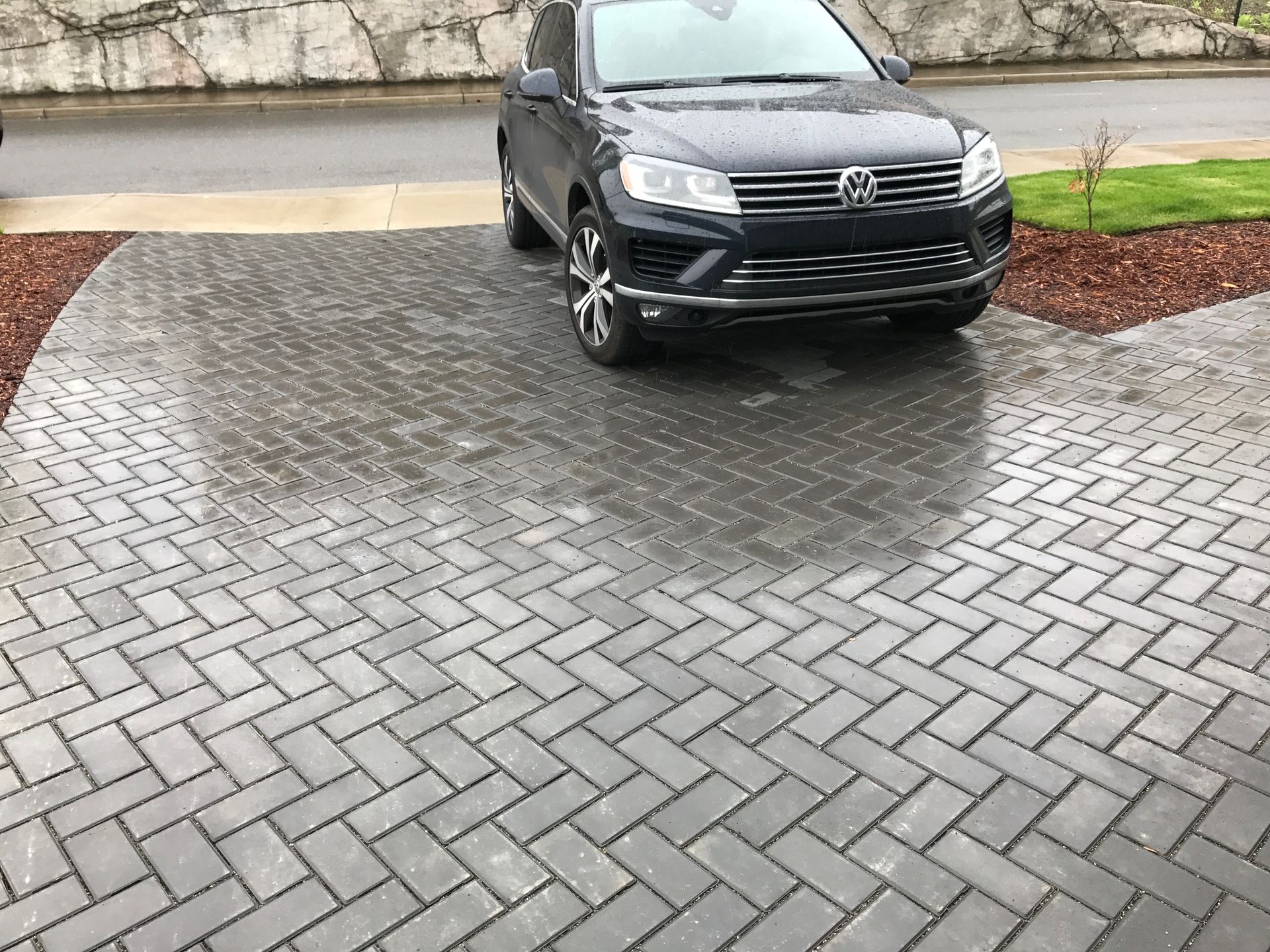 Black SUV parked on a wet gray paver driveway in front of a stone wall.
