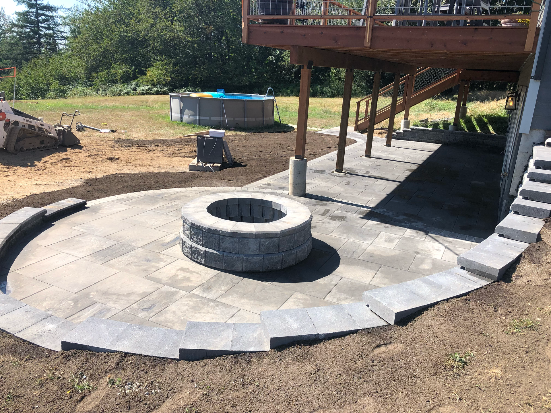 Backyard stone patio with a circular fire pit under a raised deck, surrounded by bare dirt and landscaping blocks