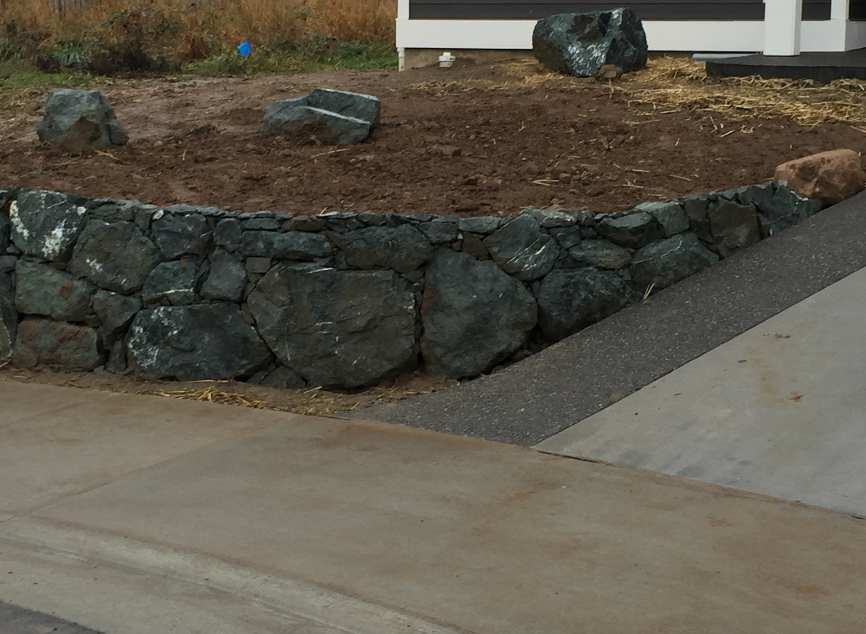 Rock retaining wall beside a sloped driveway and brown landscaped yard near a house