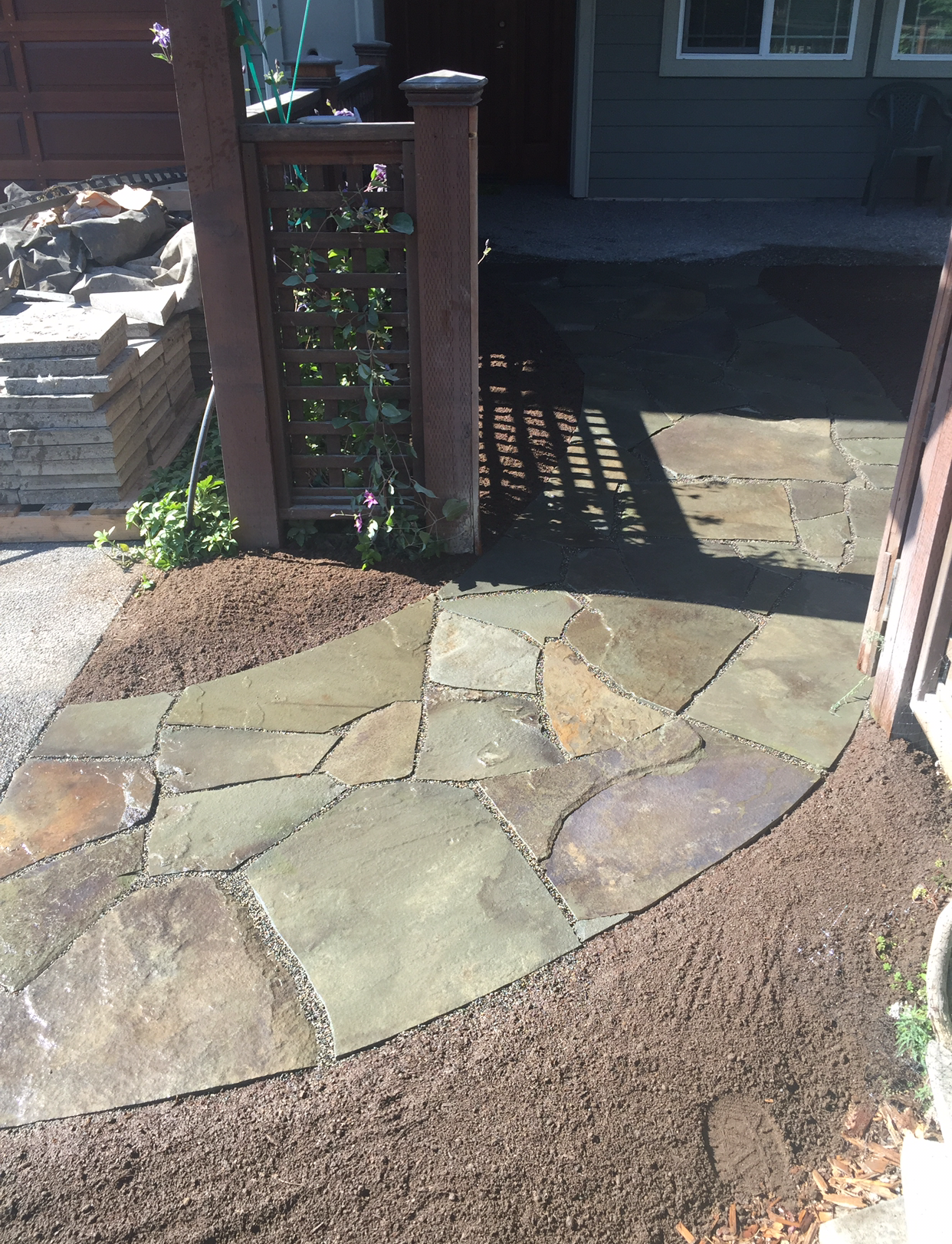 Stone walkway leading through a gate into a shaded front yard, bordered by brown gravel and a wooden fence.