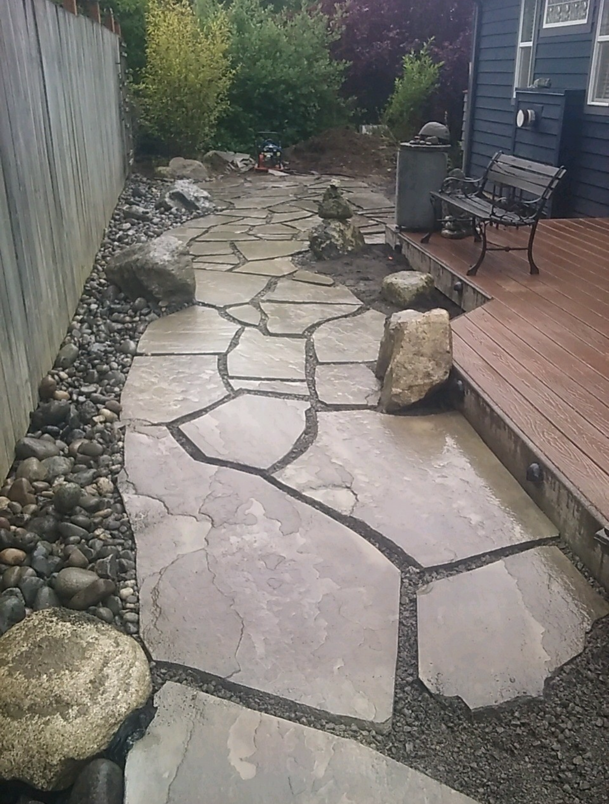 Stone garden path beside a wooden deck and fence, bordered by rocks and leading to greenery.