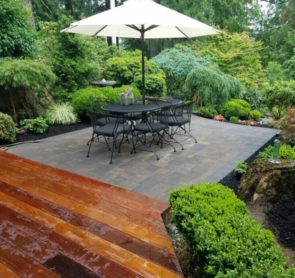 Garden patio with black metal table and chairs under a white umbrella, surrounded by lush greenery.