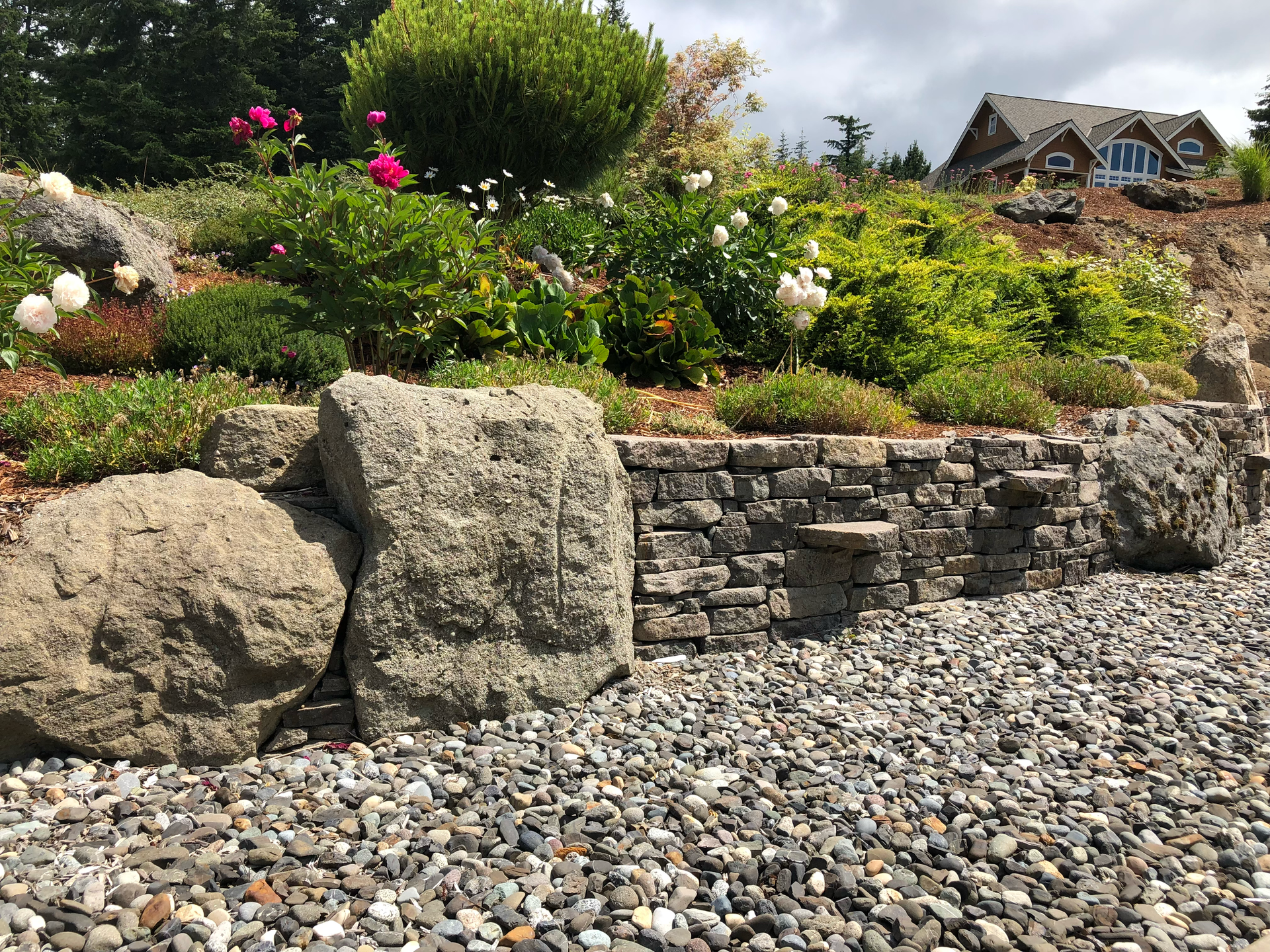Rock garden with stone retaining wall, colorful flowers, and a house in the background