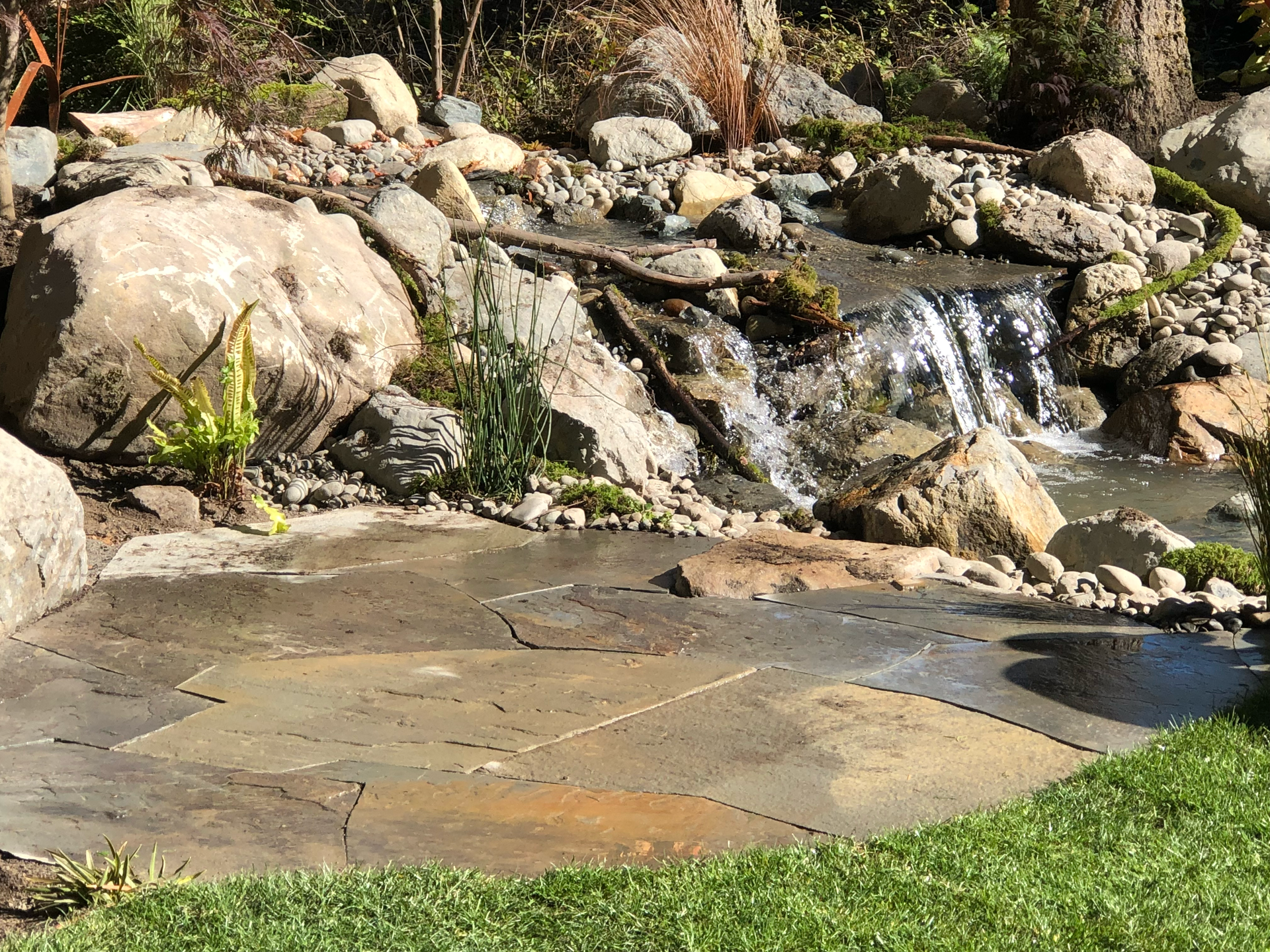 Stone patio beside a small waterfall and pond, bordered by rocks and green grass.