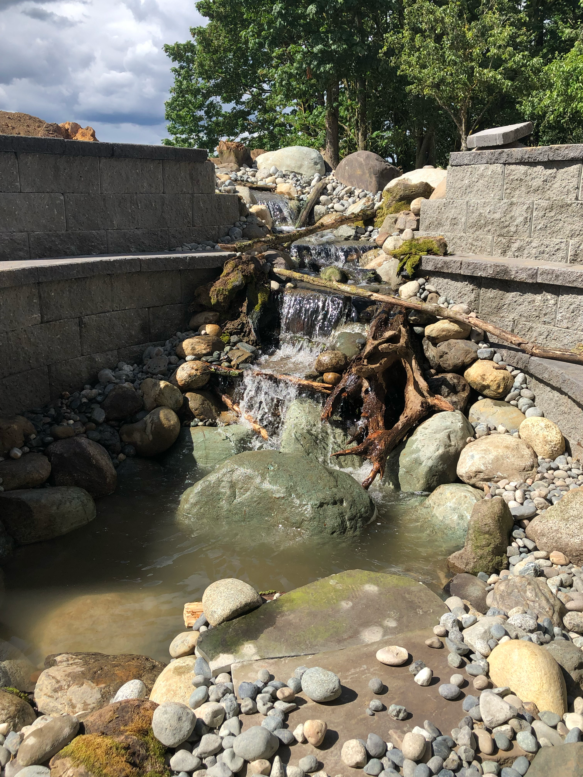 Small waterfall flowing through stone walls into a shallow rocky pool outdoors.
