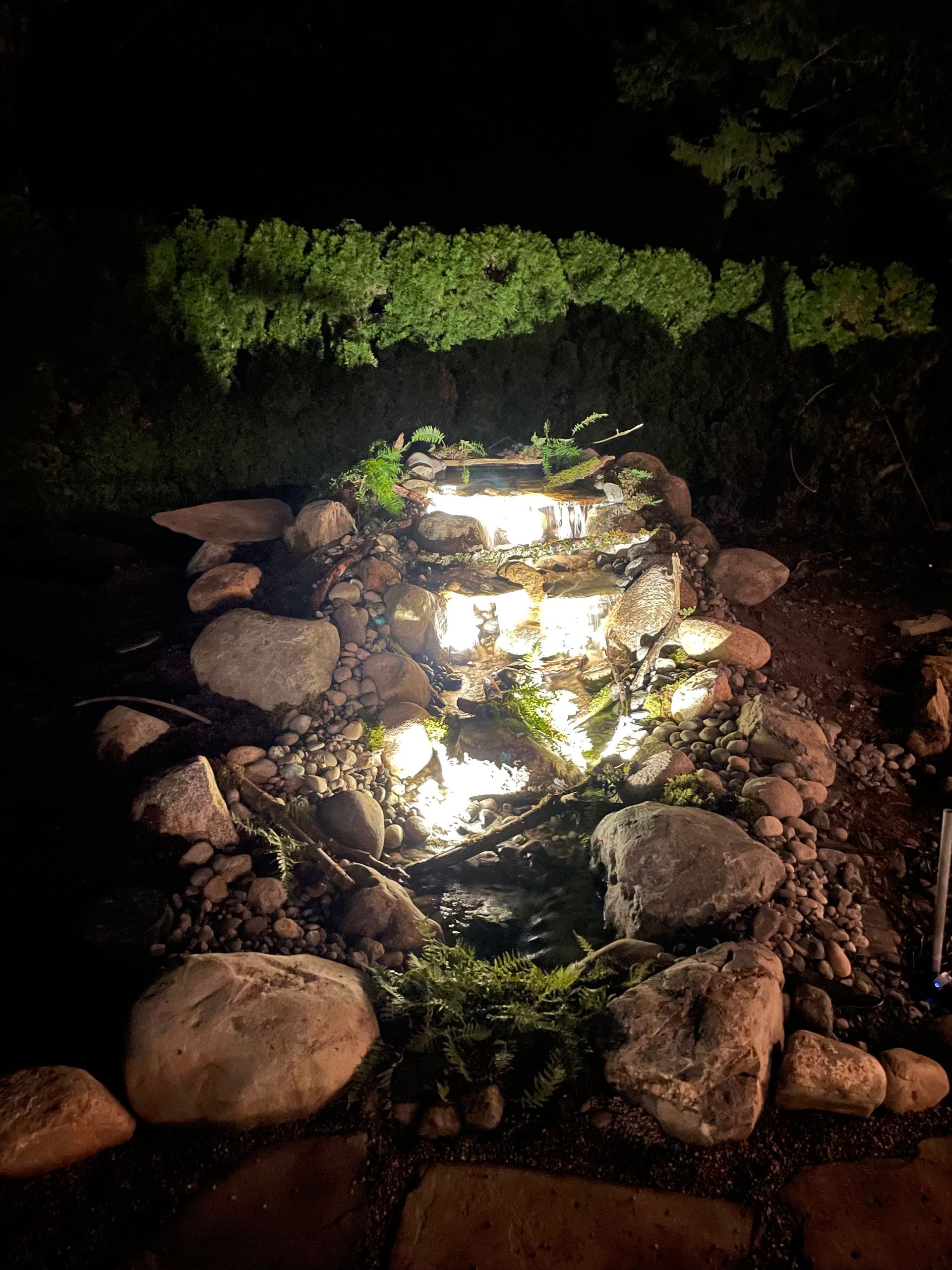 Lit garden pond with glowing water, surrounded by rocks and moss at night