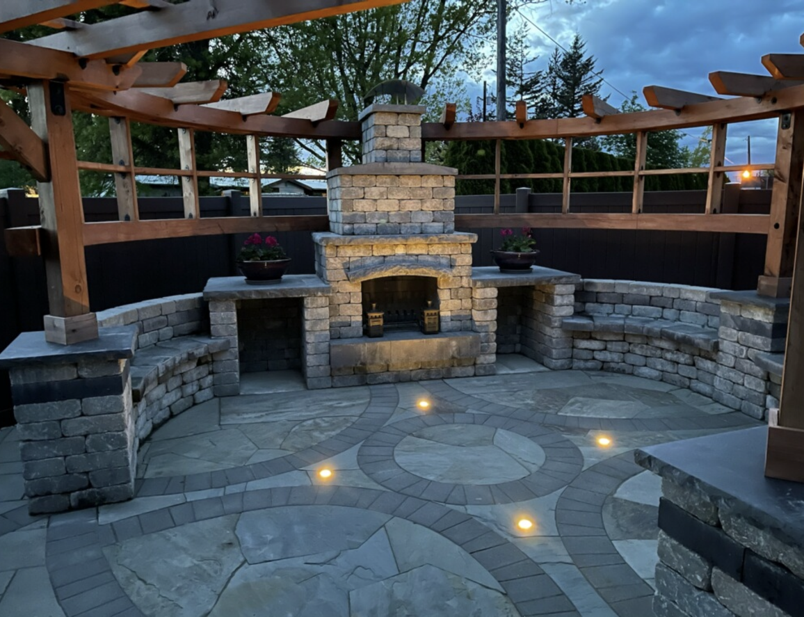 Stone outdoor patio with built-in fireplace, curved seating walls, and warm ground lights at dusk