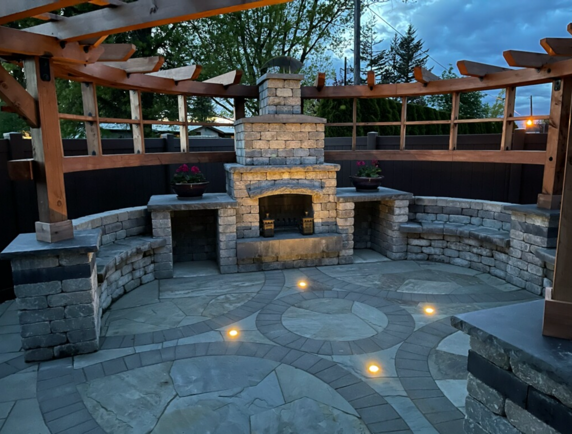 Stone outdoor fireplace and seating area under a pergola at dusk, with a circular flagstone patio and warm lights.