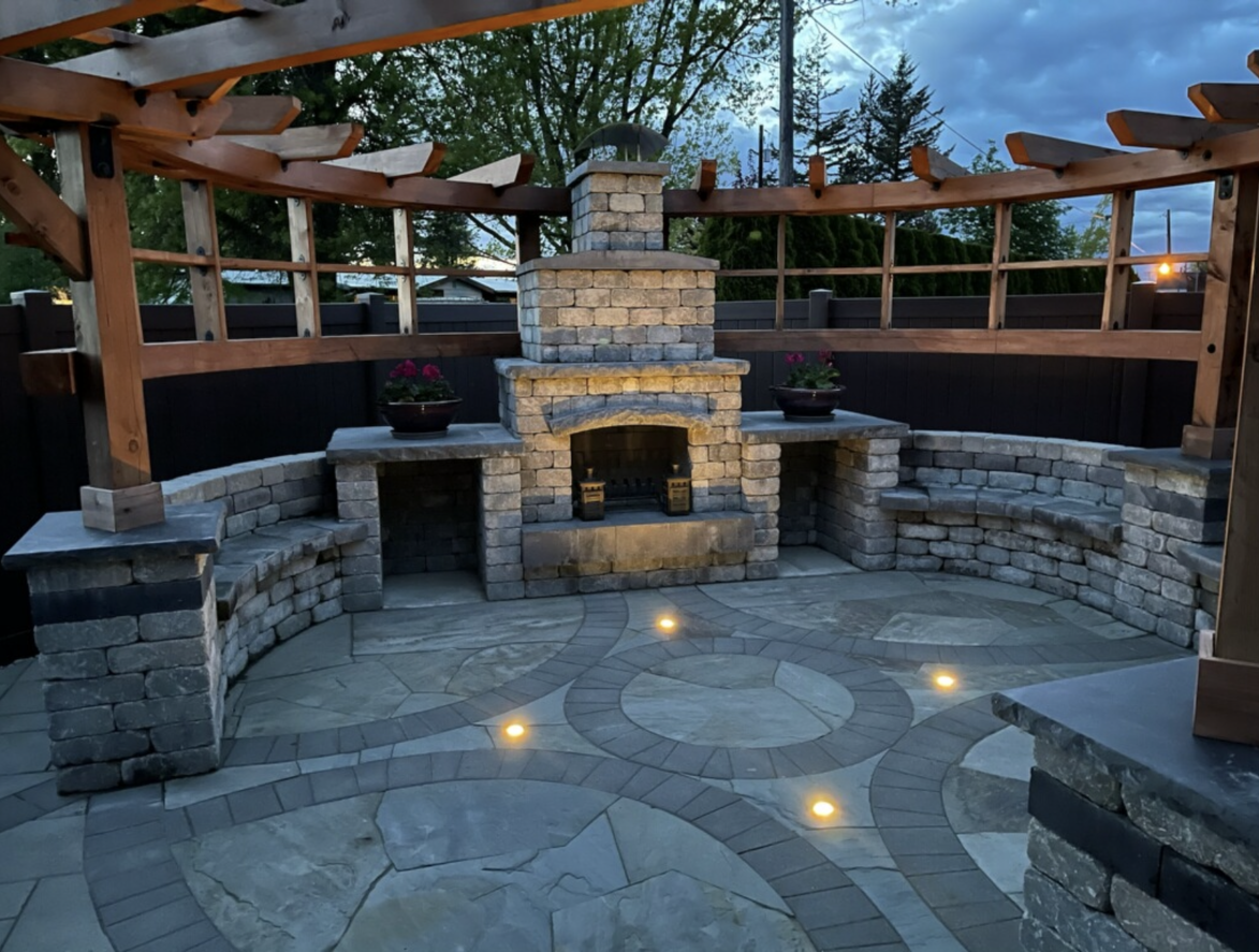 Stone patio with a built-in fireplace under a wooden pergola at dusk