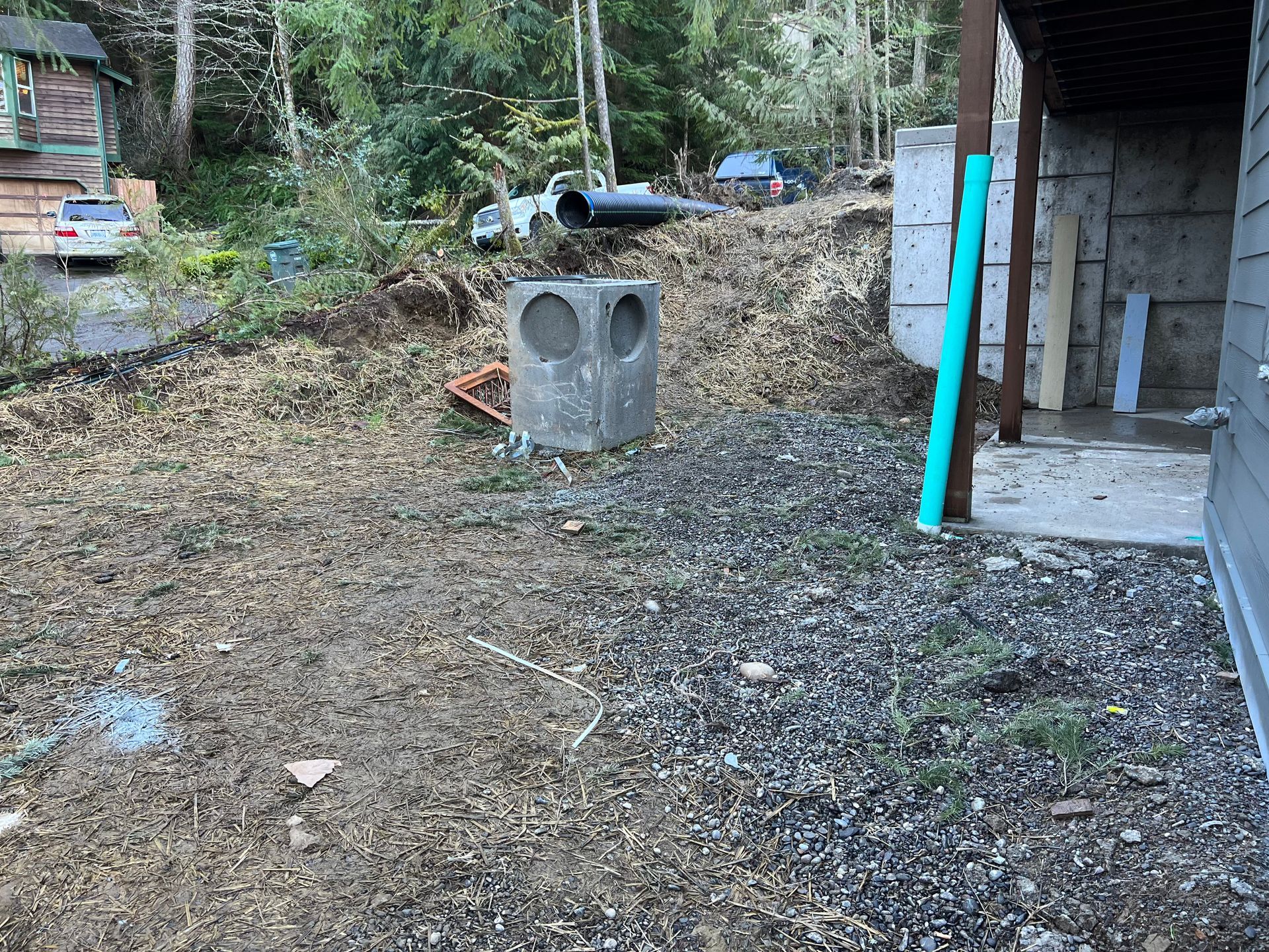 Backyard with a dirt-and-gravel slope, concrete structure, blue pipe, and wooded hillside in the background