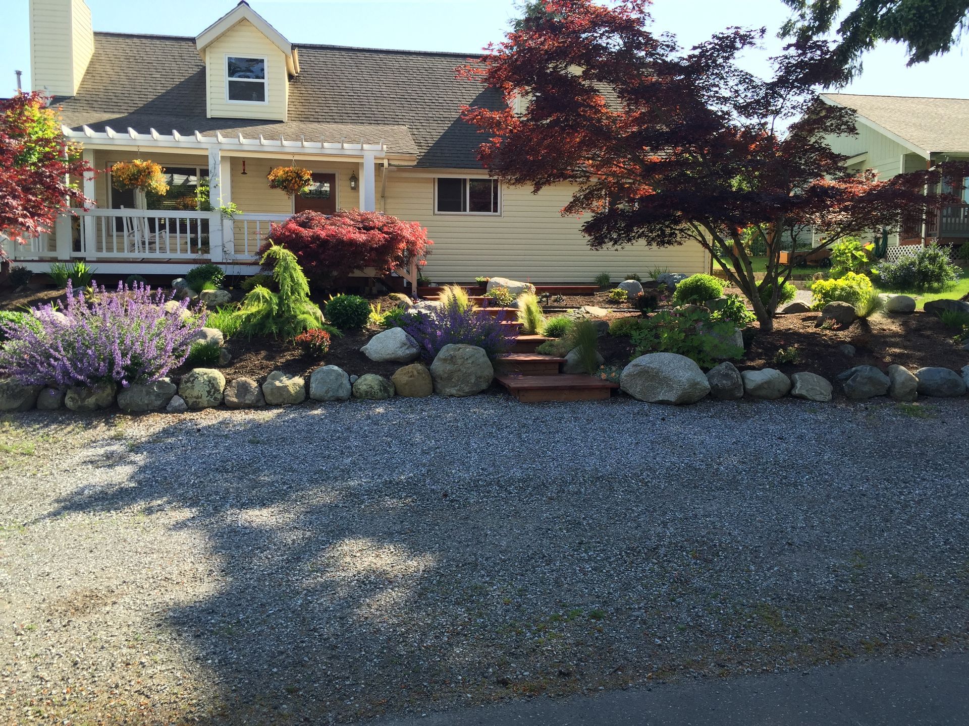 House with porch, landscaped garden, stone border, and gravel driveway under sunny daylight