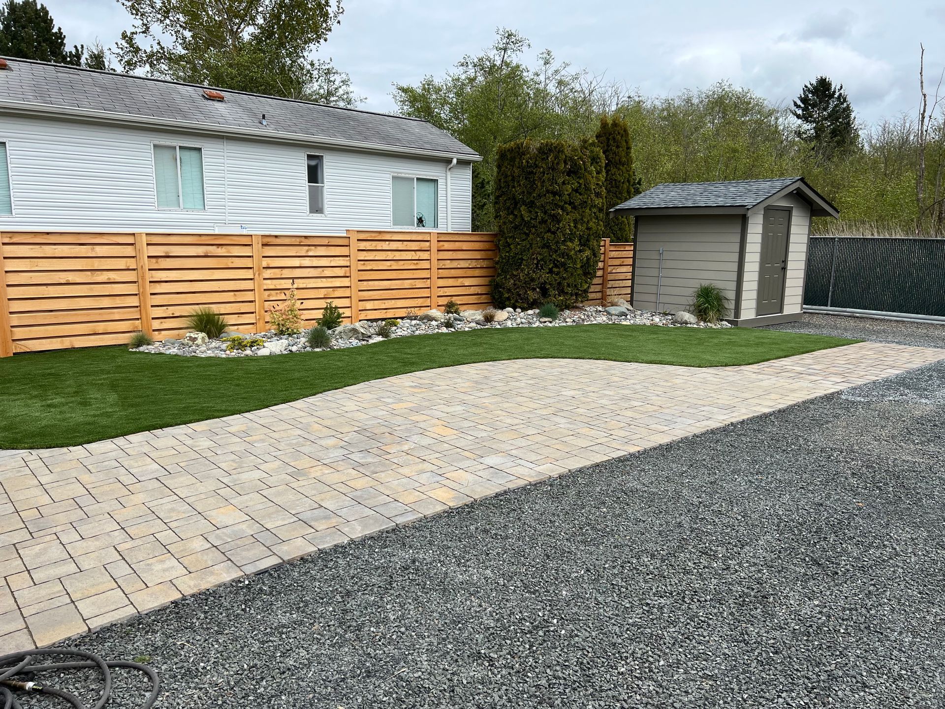 Backyard with a stone path, gravel driveway, green lawn, wooden fence, and small shed.