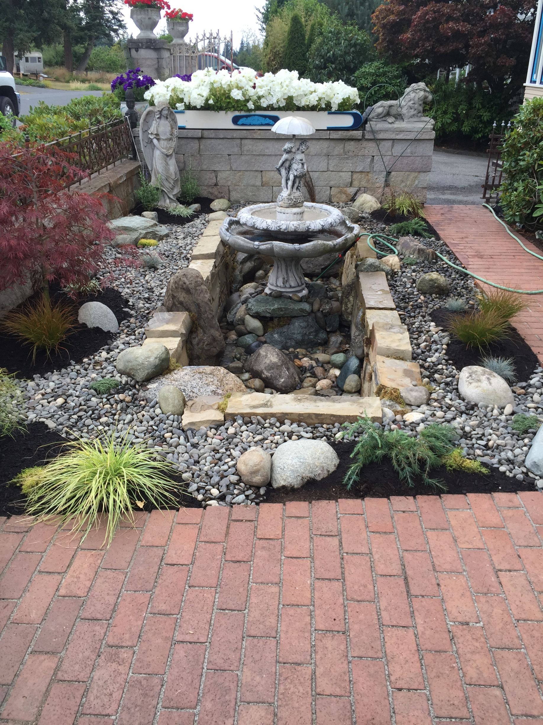 Small garden fountain surrounded by rocks and flowers, bordered by a red brick walkway.