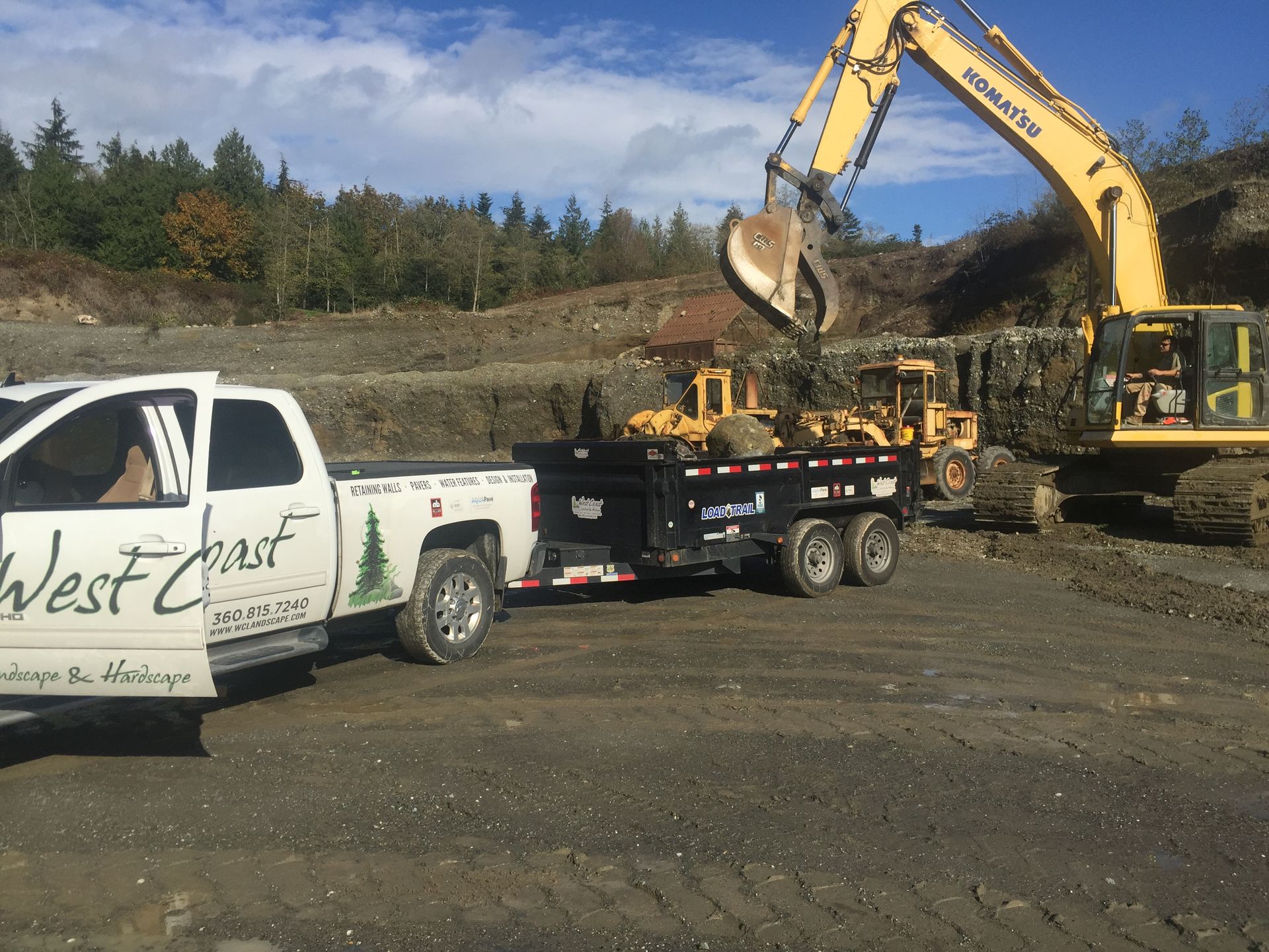 White pickup with trailer and excavator at a dirt construction site under clear sky