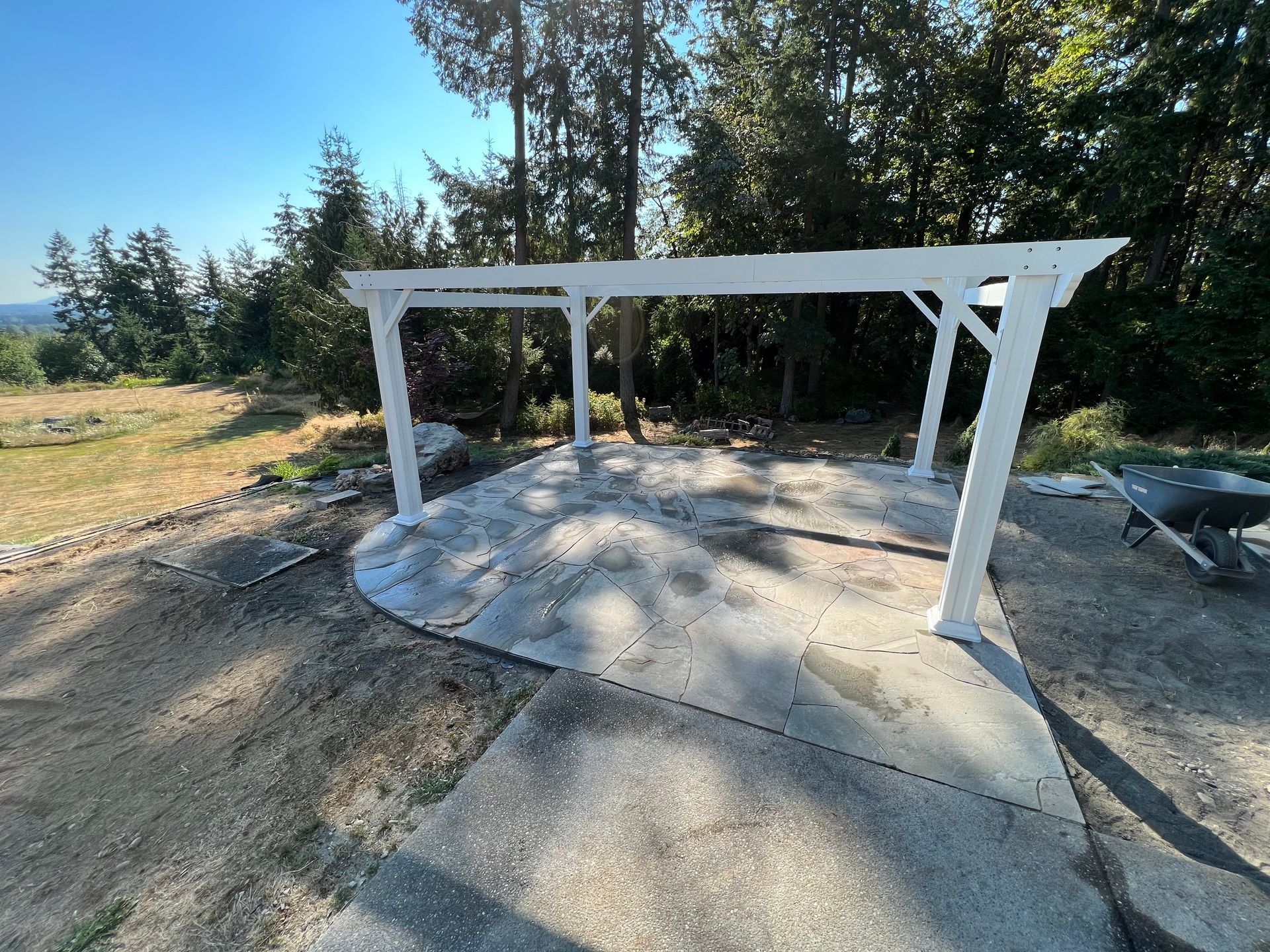 White pergola structure over a concrete pad in a wooded outdoor area, with a wheelbarrow nearby.
