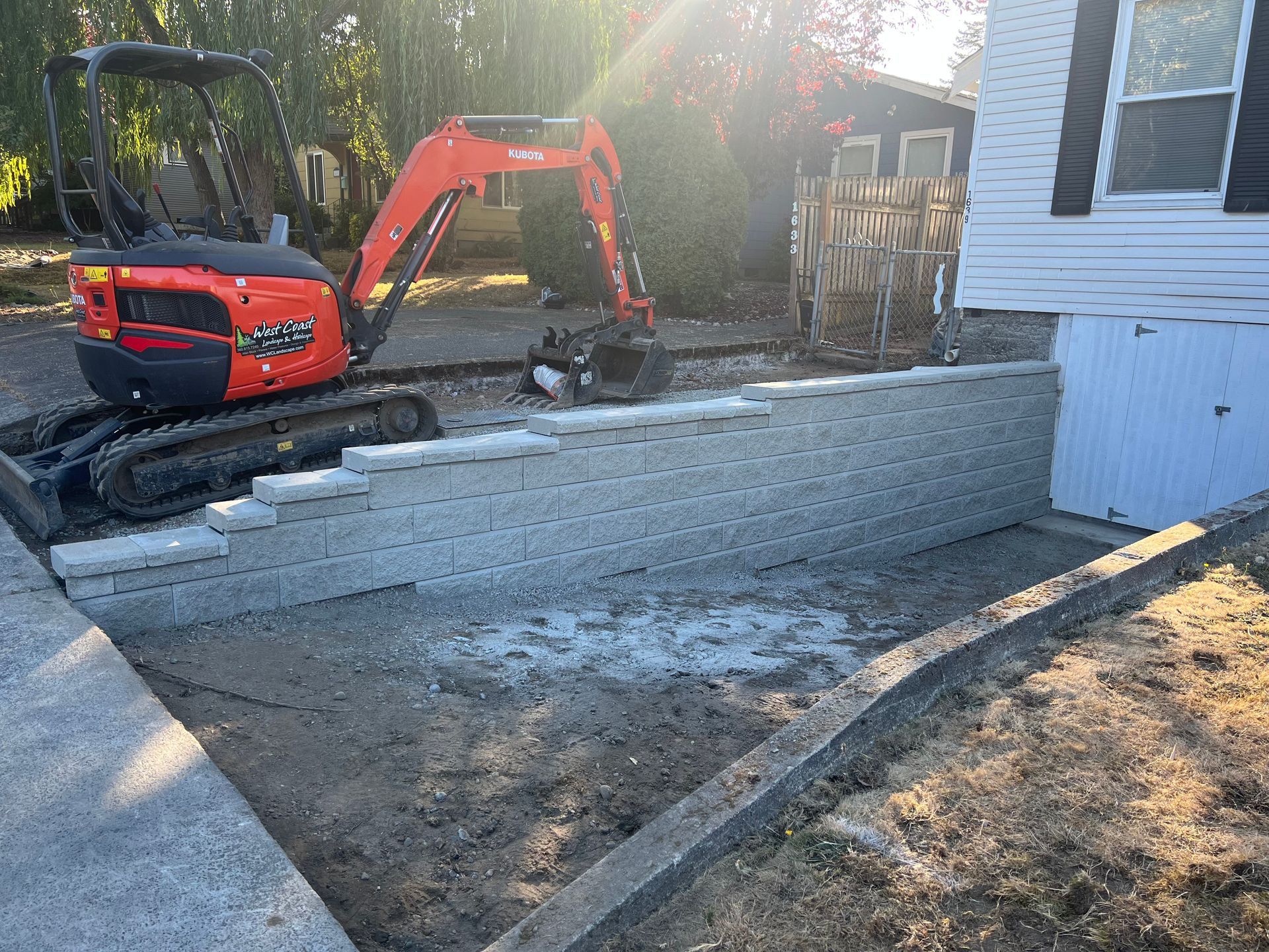 Backhoe and worker building a concrete retaining wall beside a house, with steps and dirt grading.