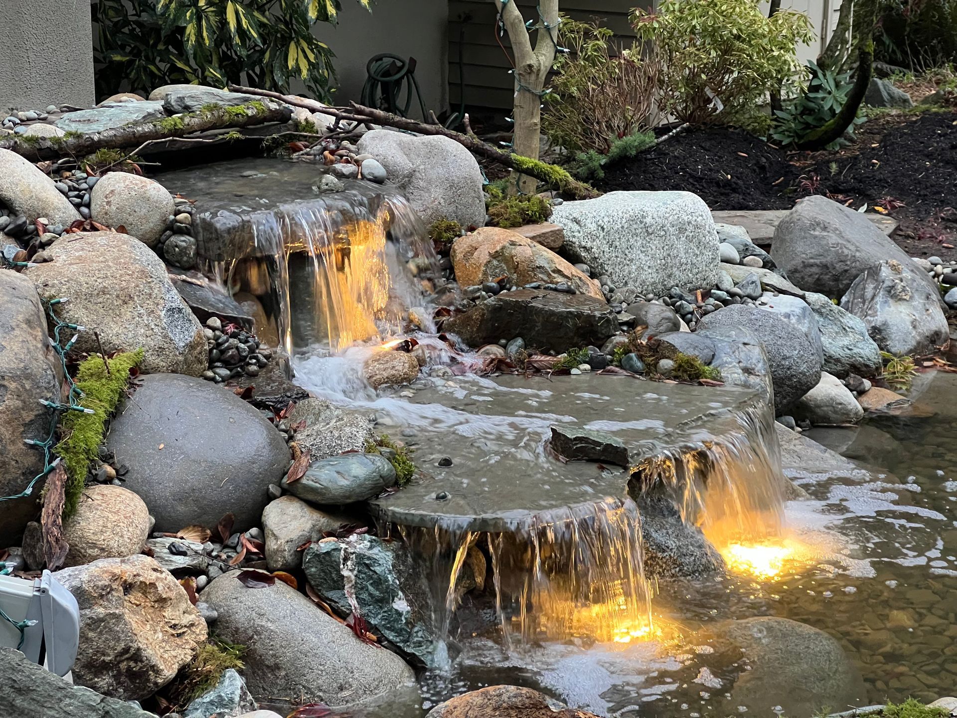 Backyard rock waterfall with flowing water and glowing lights among stones and greenery