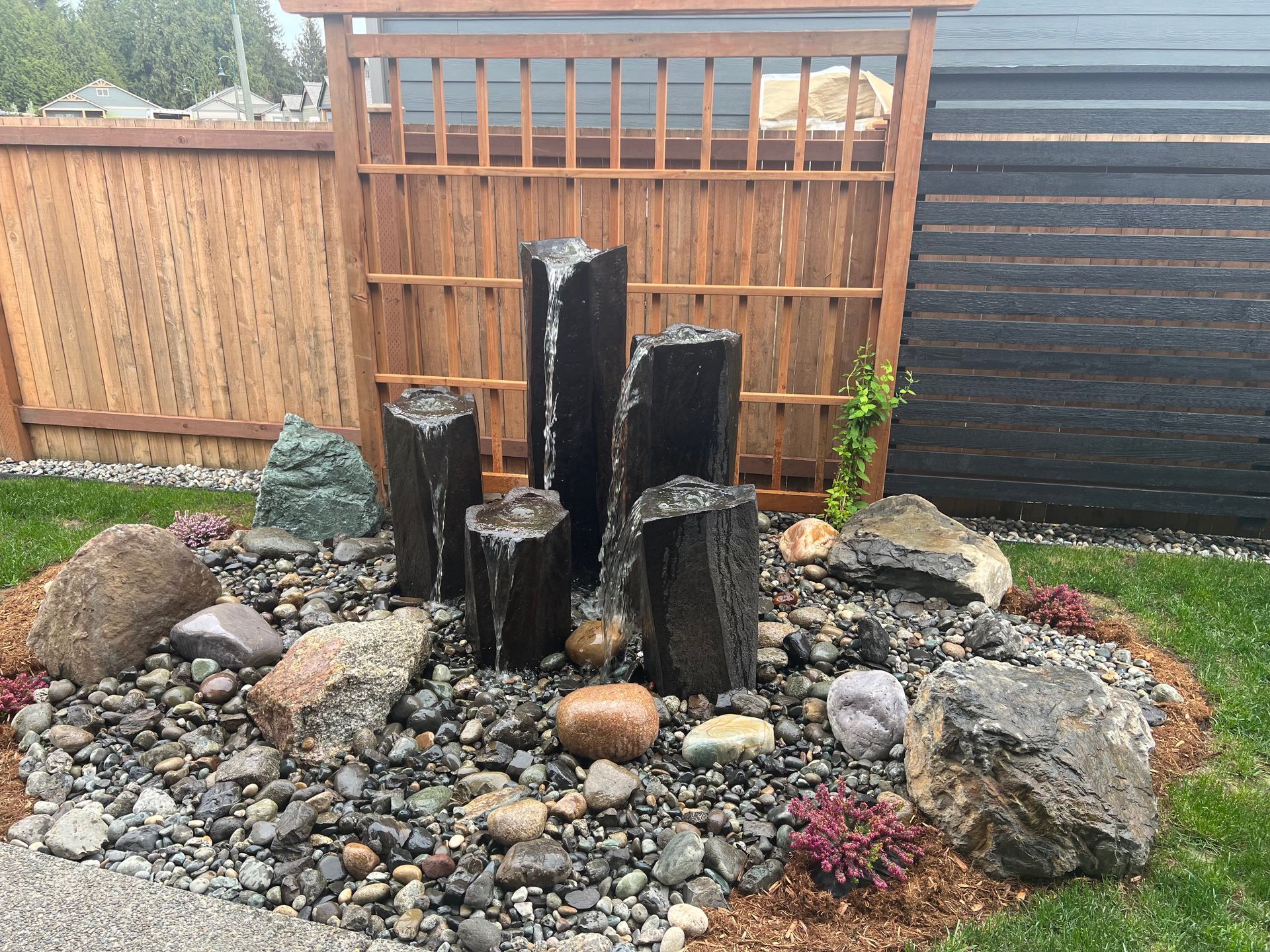 Small backyard rock fountain with black stone columns, surrounded by pebbles, plants, and a wooden fence.