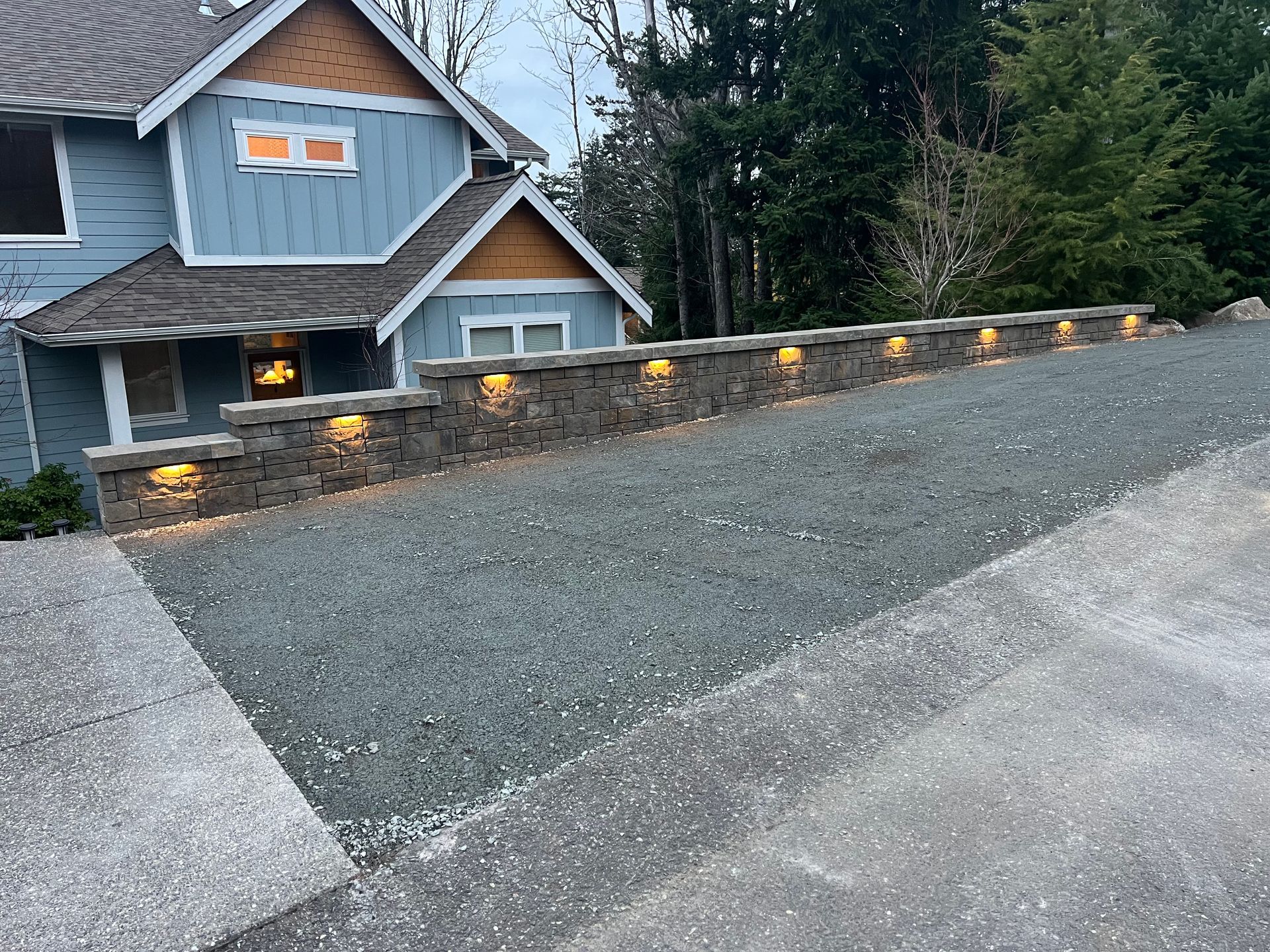 Sloped gravel driveway beside a blue house with lit stone wall lights at dusk