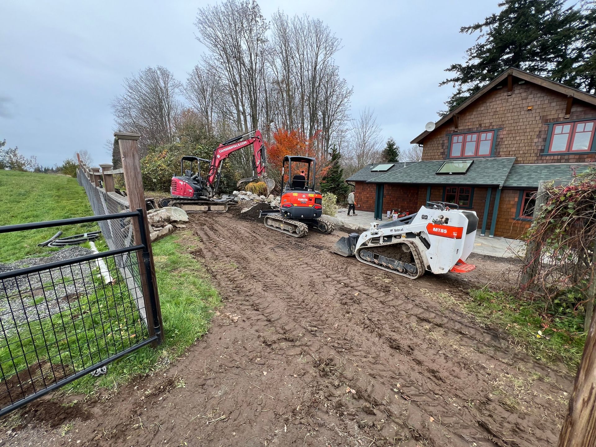 Excavator and skid steer working on muddy driveway beside a brick house