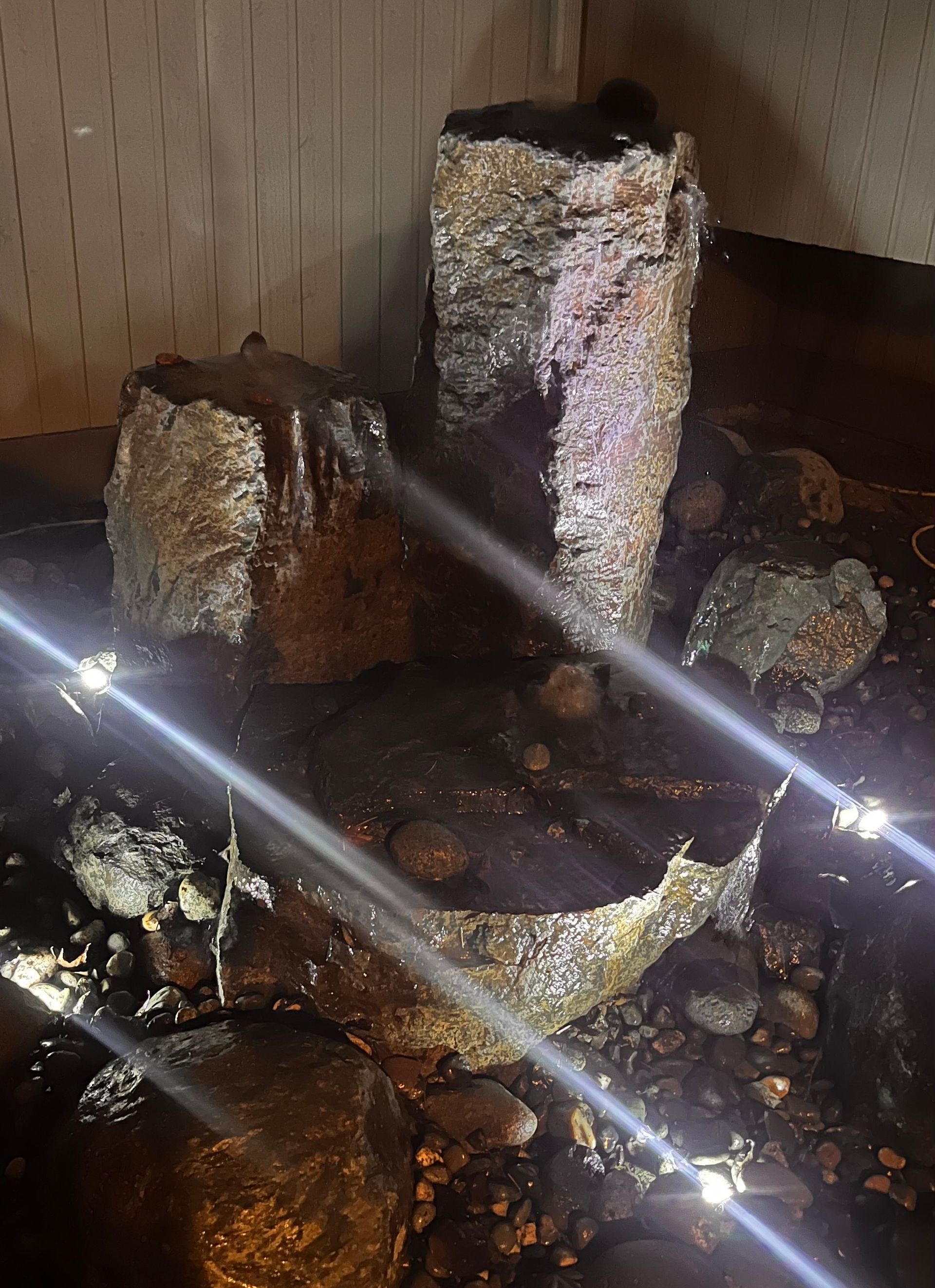 Indoor decorative water fountain with rocks and a central stone spout, lit by bright reflections.