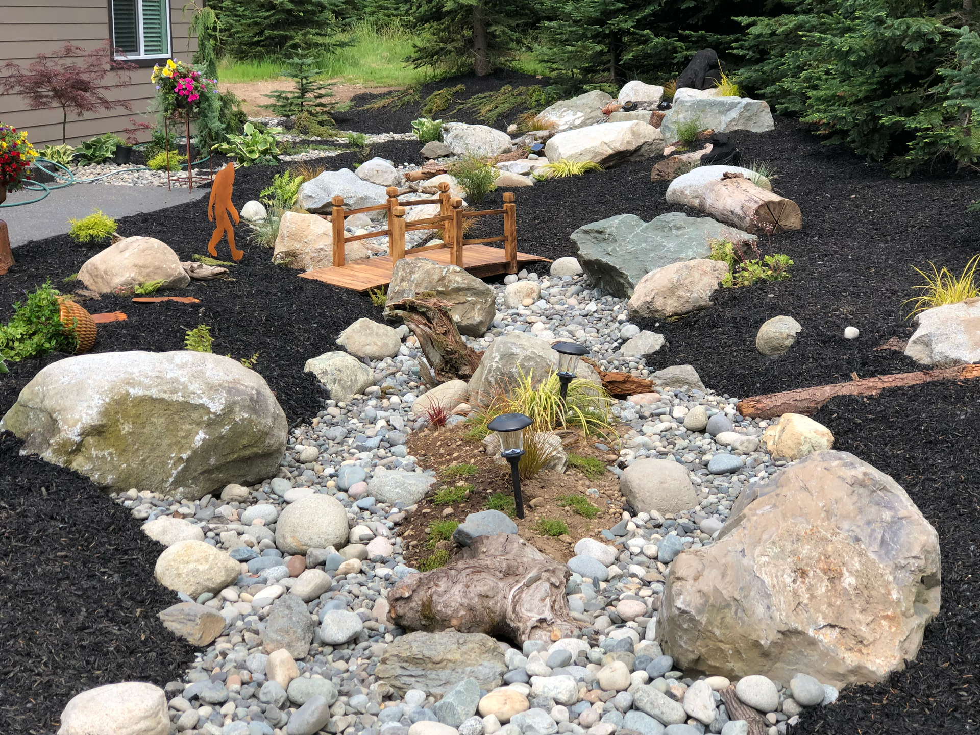 Rock garden with large stones, white pebbles, and a small wooden bridge in a landscaped yard.