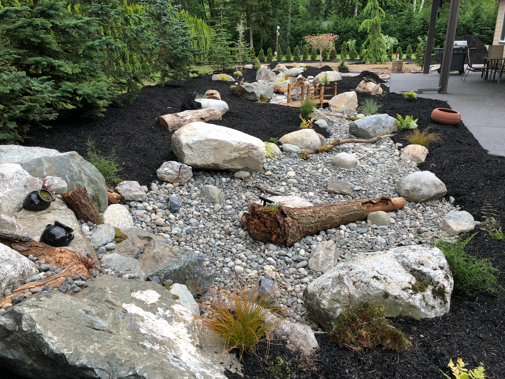 Rock garden with large boulders, gravel, and scattered logs beside green shrubs and trees