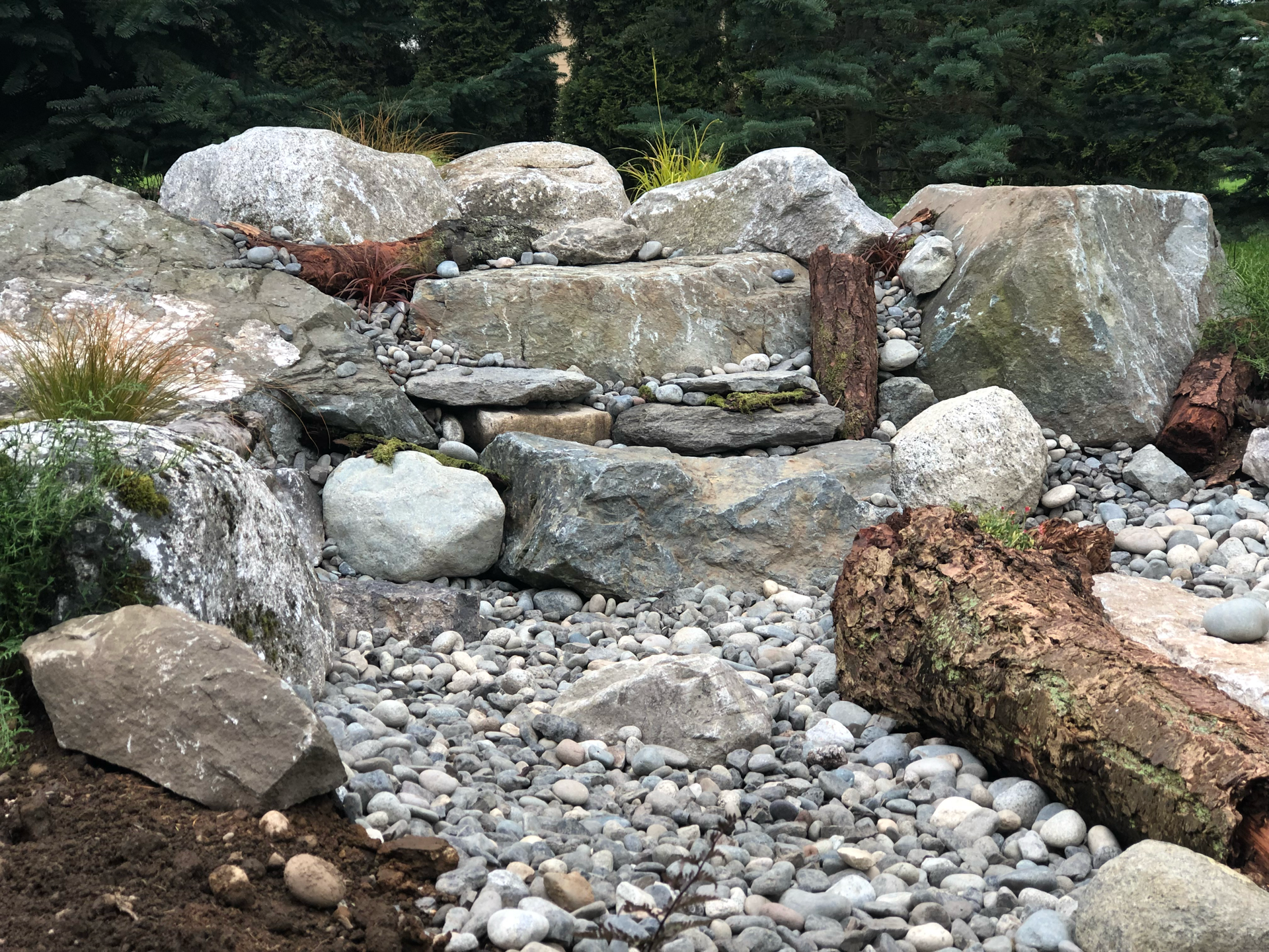 Rock garden with stacked stones, pebbles, and logs in front of greenery.